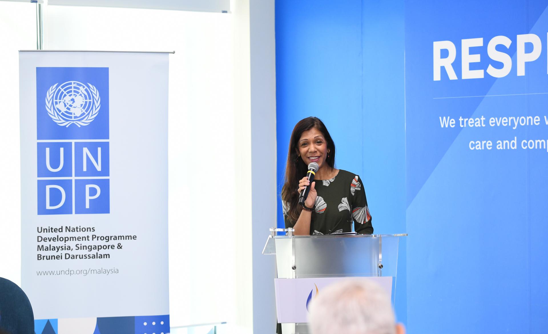 A woman speaking into a microphone at a podium with a UNDP banner in a bright room.