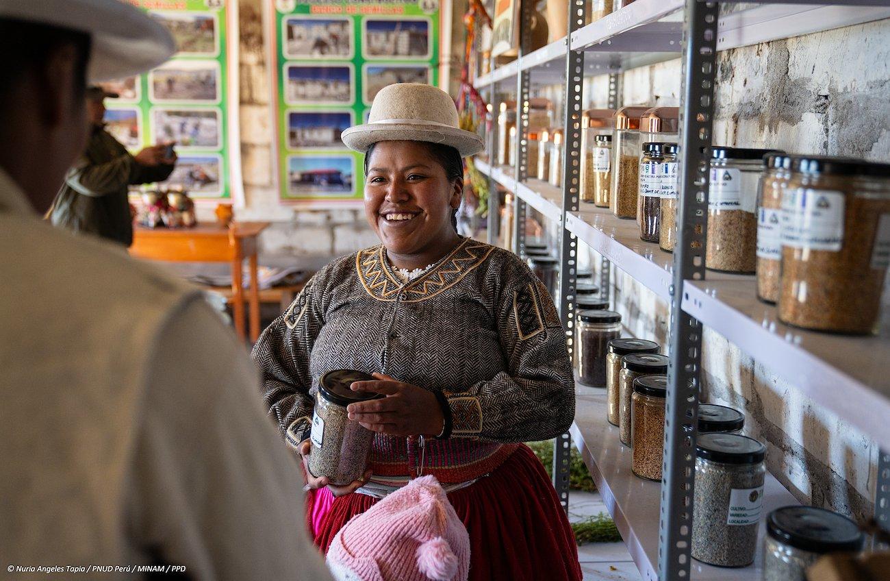 A smiling woman in traditional attire stands in a colorful shop filled with jars of spices.