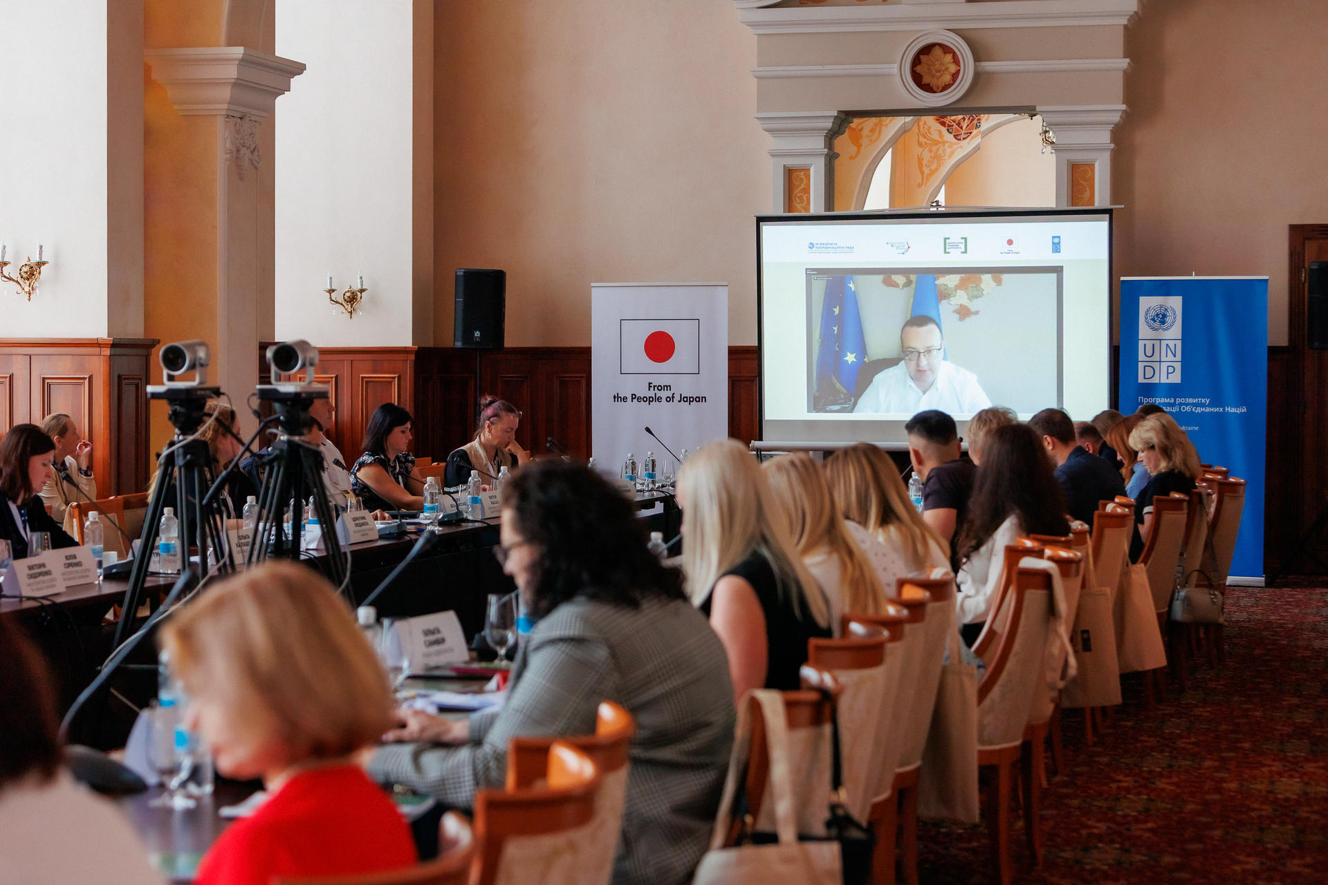 A conference setting with attendees seated at long tables, focused on a presenter on screen.
