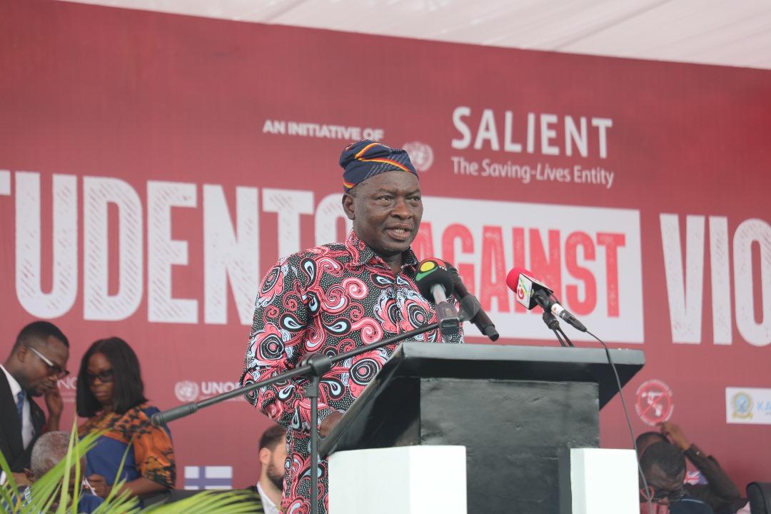 A man in traditional attire speaks at a podium, with a "Students Against Violence" backdrop.