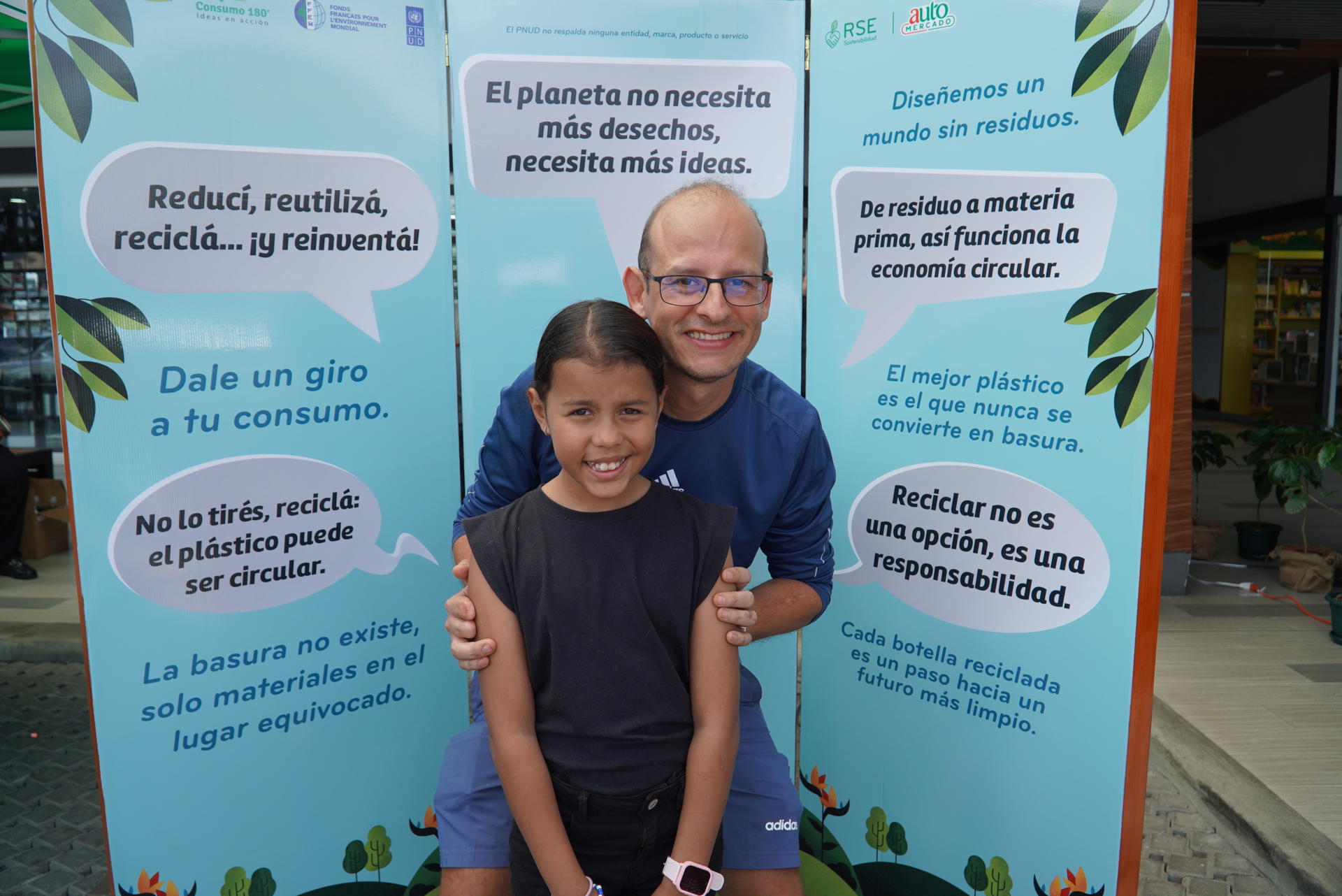 A man and a girl pose together, smiling, in front of colorful banners promoting recycling.