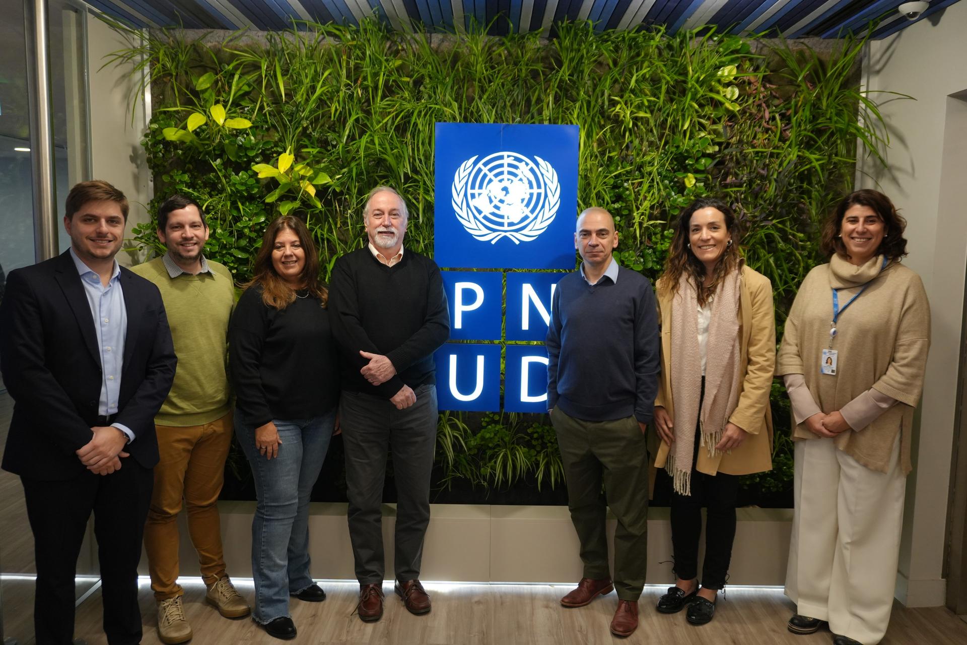 Un grupo de seis personas posando frente a una pared con plantas verdes y el logo de PNUD.