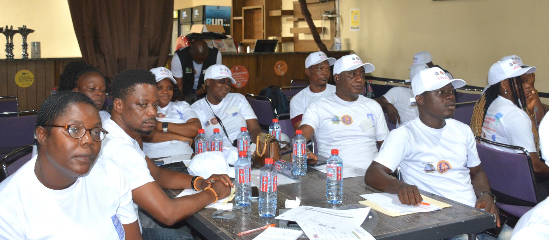A group of individuals wearing white shirts and caps are seated at tables, attentively listening.