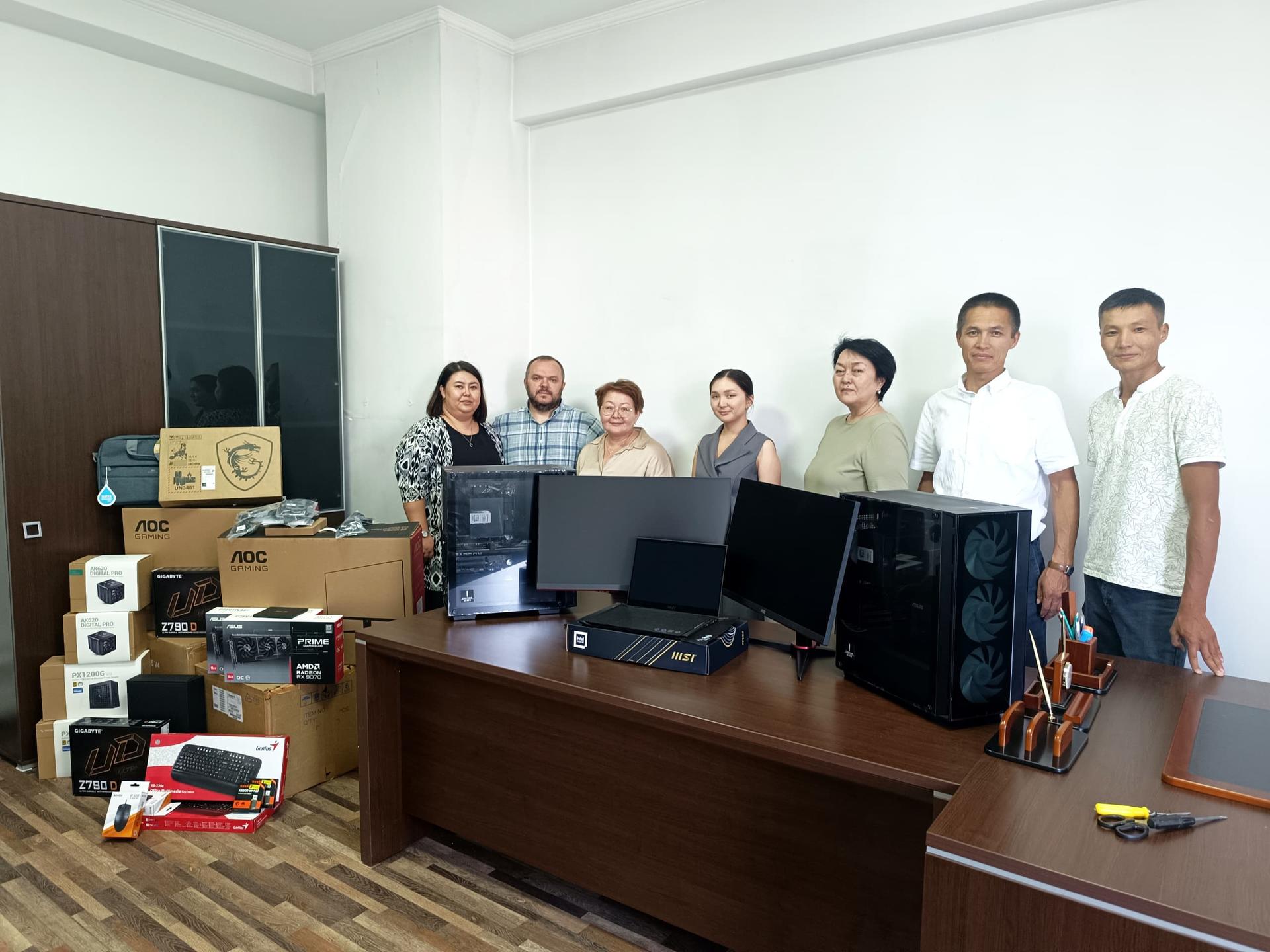 A group of six people stands in an office among computer equipment and boxes.