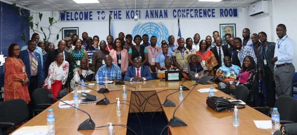 Diverse group of people gathered in a conference room with a table and UN banners.