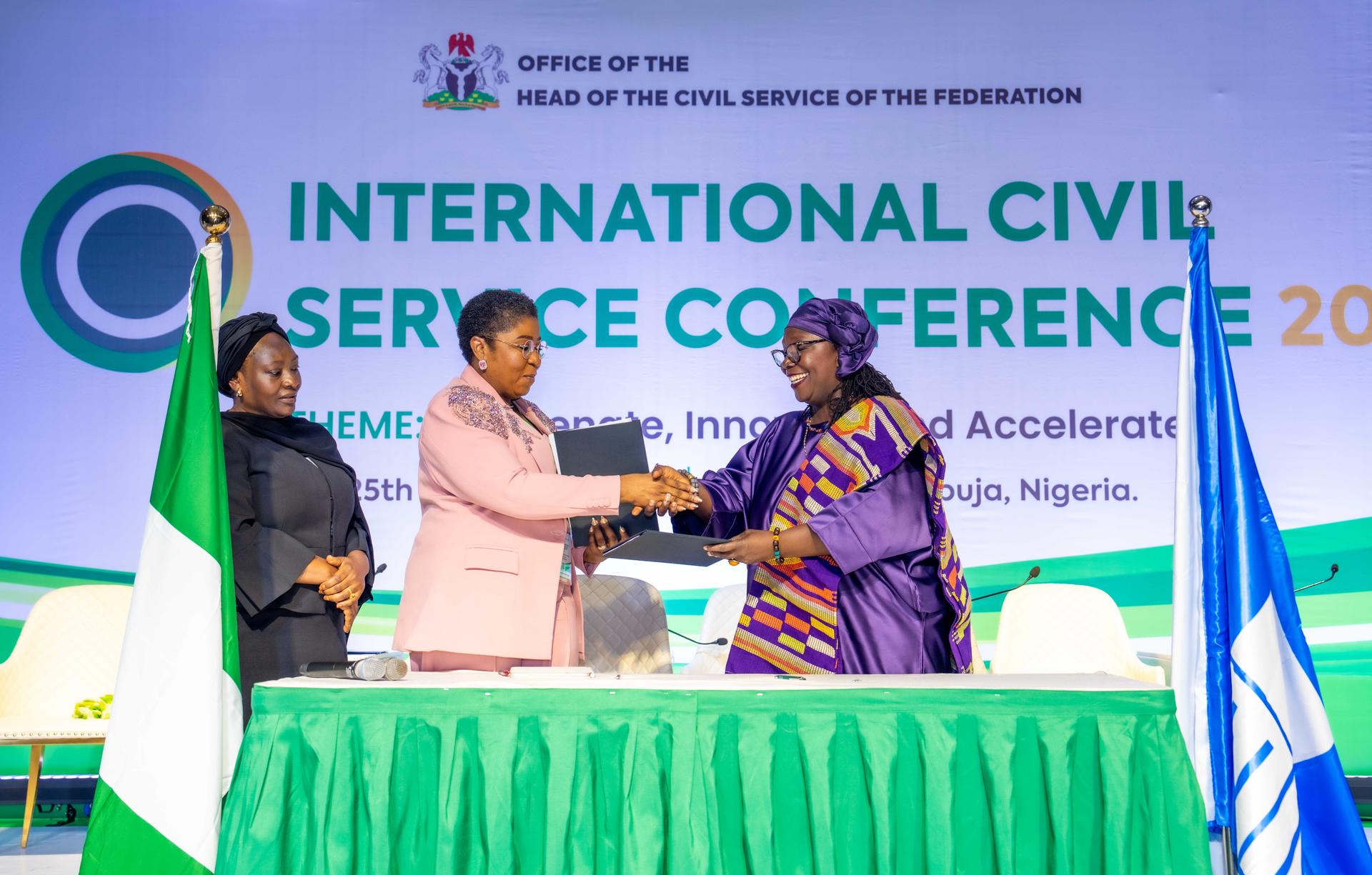 Two women shake hands at a conference table, with flags in the background and a banner above.