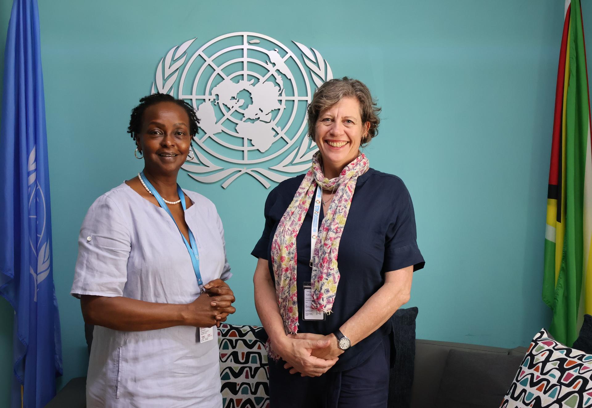 Two women stand smiling in front of the UN emblem, surrounded by flags and a cozy setting.