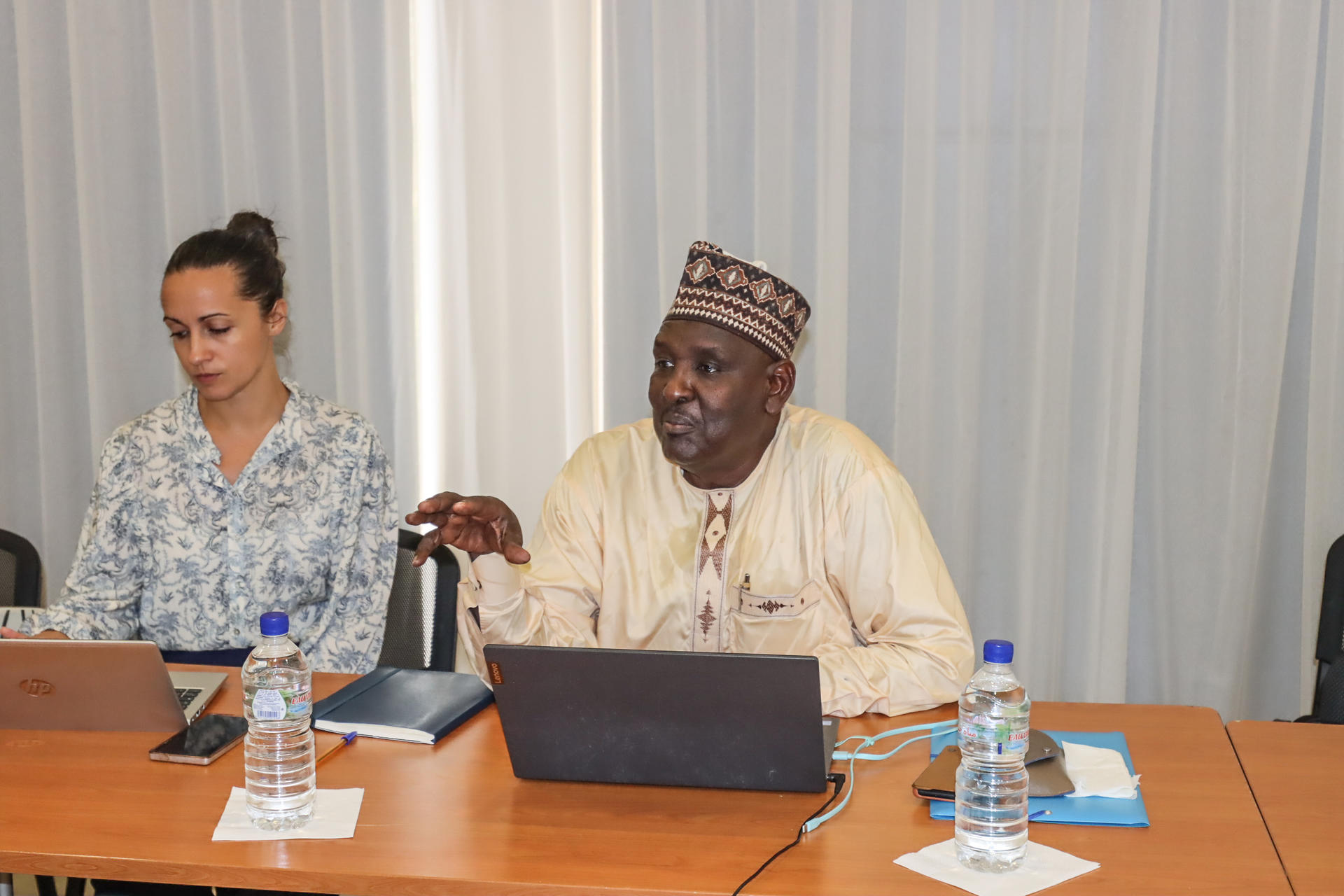 A man in traditional attire speaks at a conference table, while a woman attentively listens.