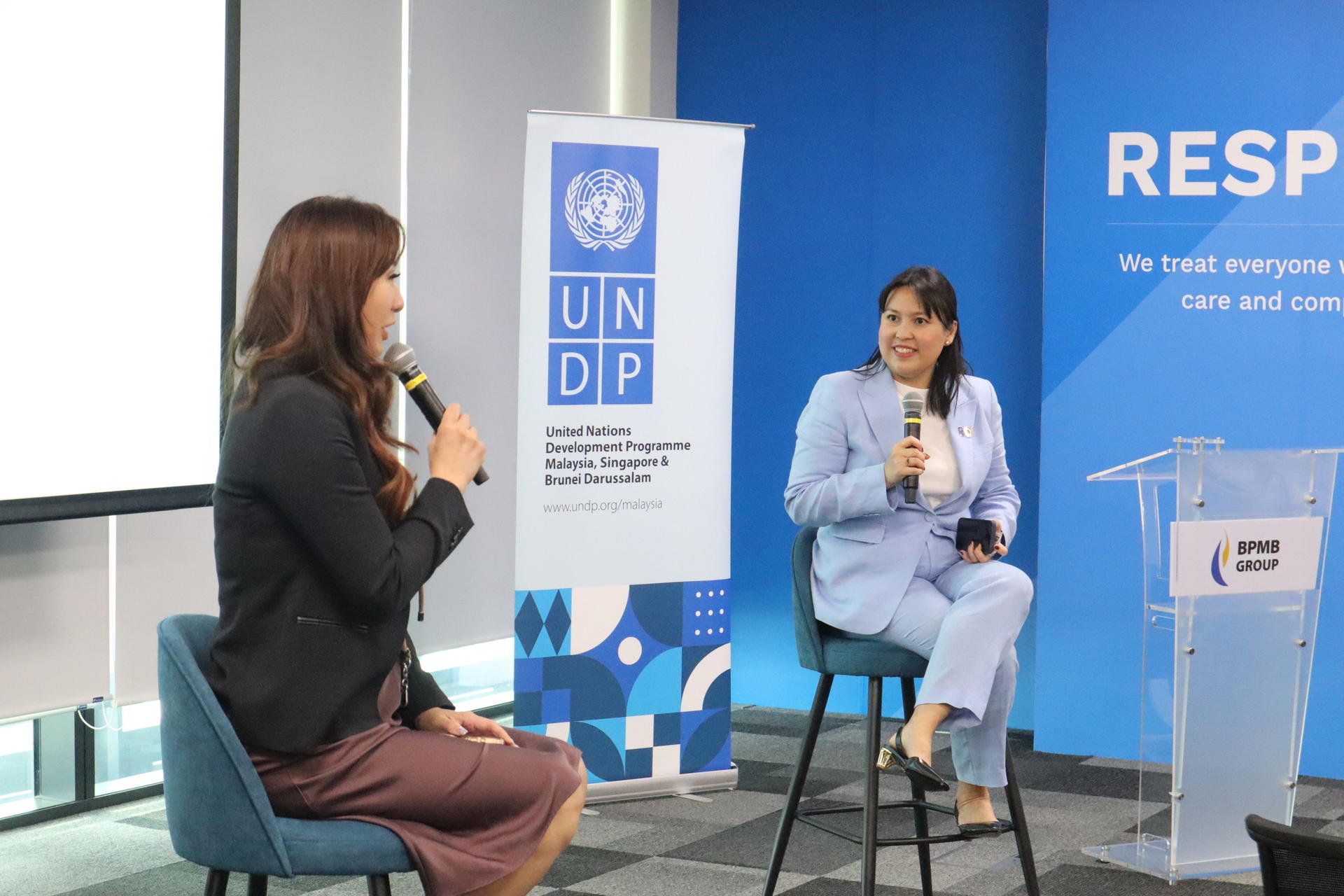 Two women speaking at a conference, seated on high stools with a UNDP banner in the background.