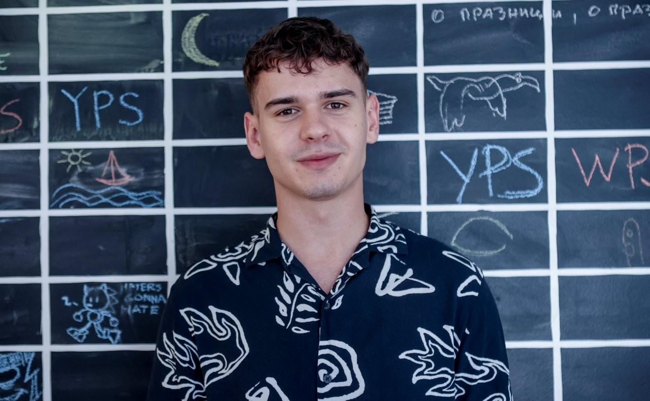Young man with curly hair smiles in front of a chalkboard with colorful drawings.