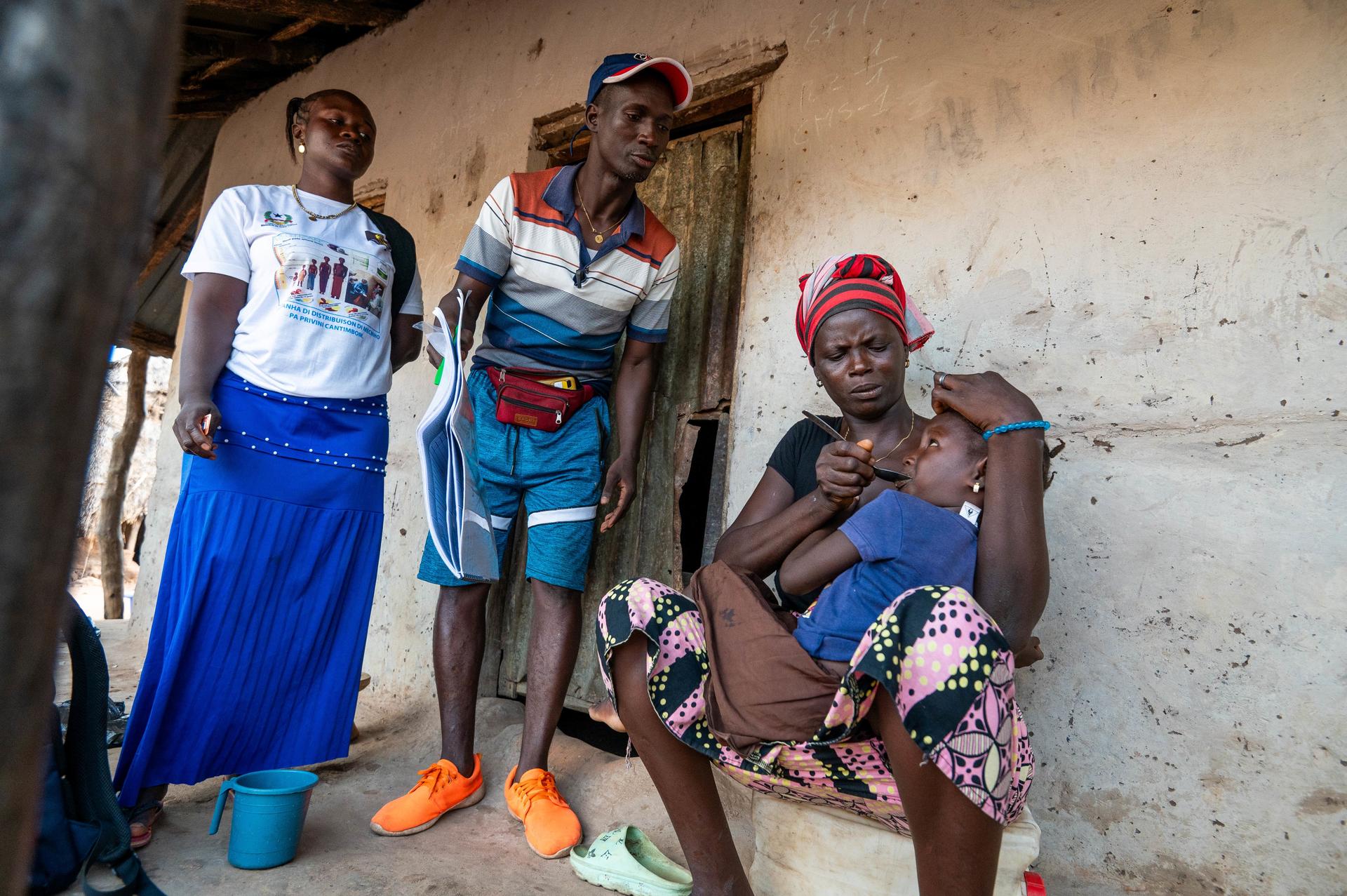 A woman holds a child on her lap, while two people stand nearby in a rural setting.