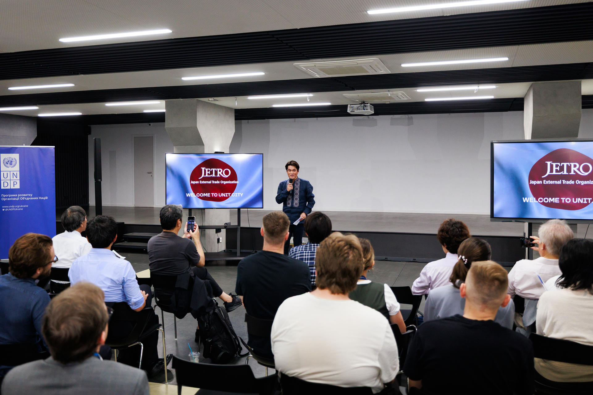 A speaker presents on stage to an audience in a modern conference setting.