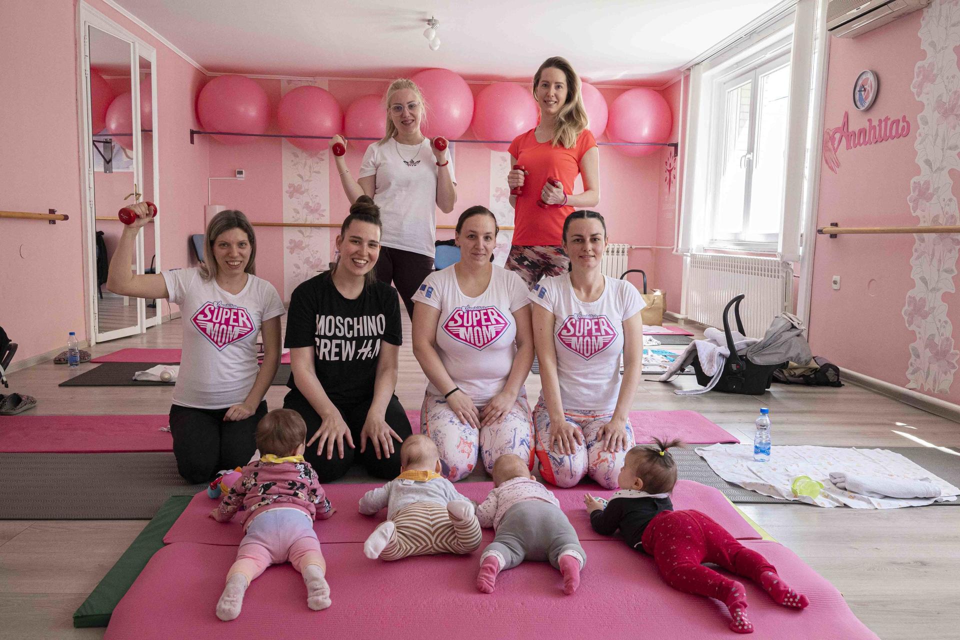 A group of women holding weights poses with babies on exercise mats in a bright pink studio.