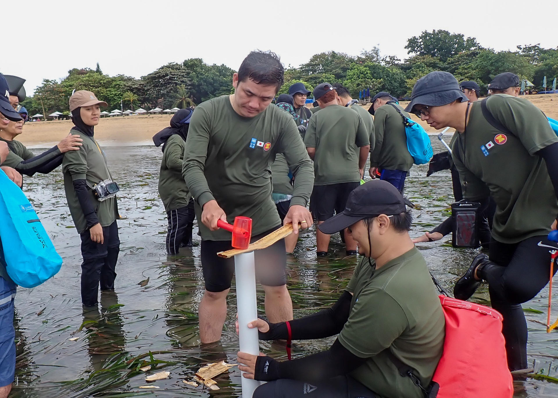 Group of people in green shirts conducting beach cleanup, using tools near the water.