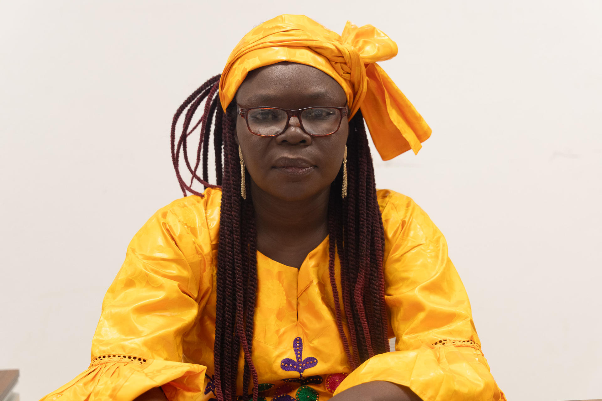 Woman wearing a vibrant yellow traditional outfit and glasses, seated and looking at the camera.