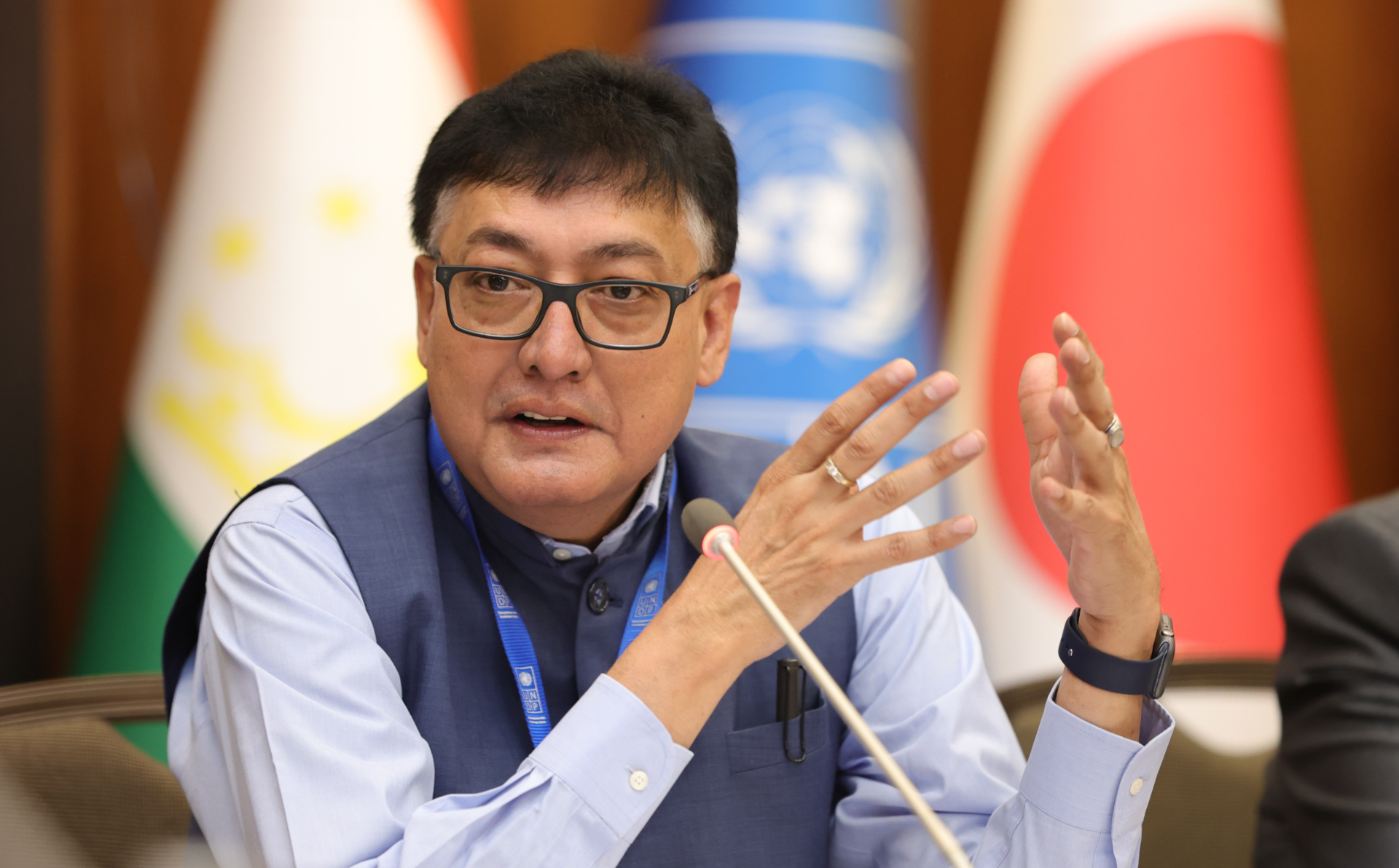 A man in glasses gestures while speaking at a conference, with flags in the background.