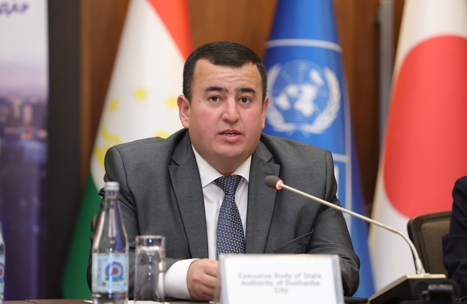 A man in a suit speaks at a conference, flags and a logo visible in the background.