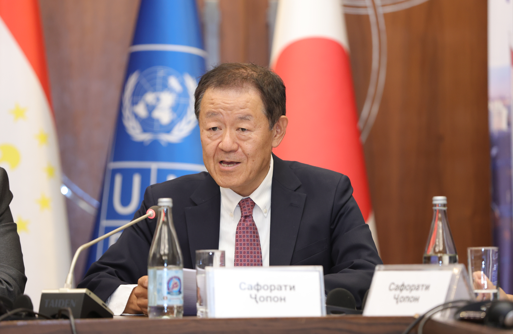 A man in a suit speaking at a conference table with flags and logos in the background.