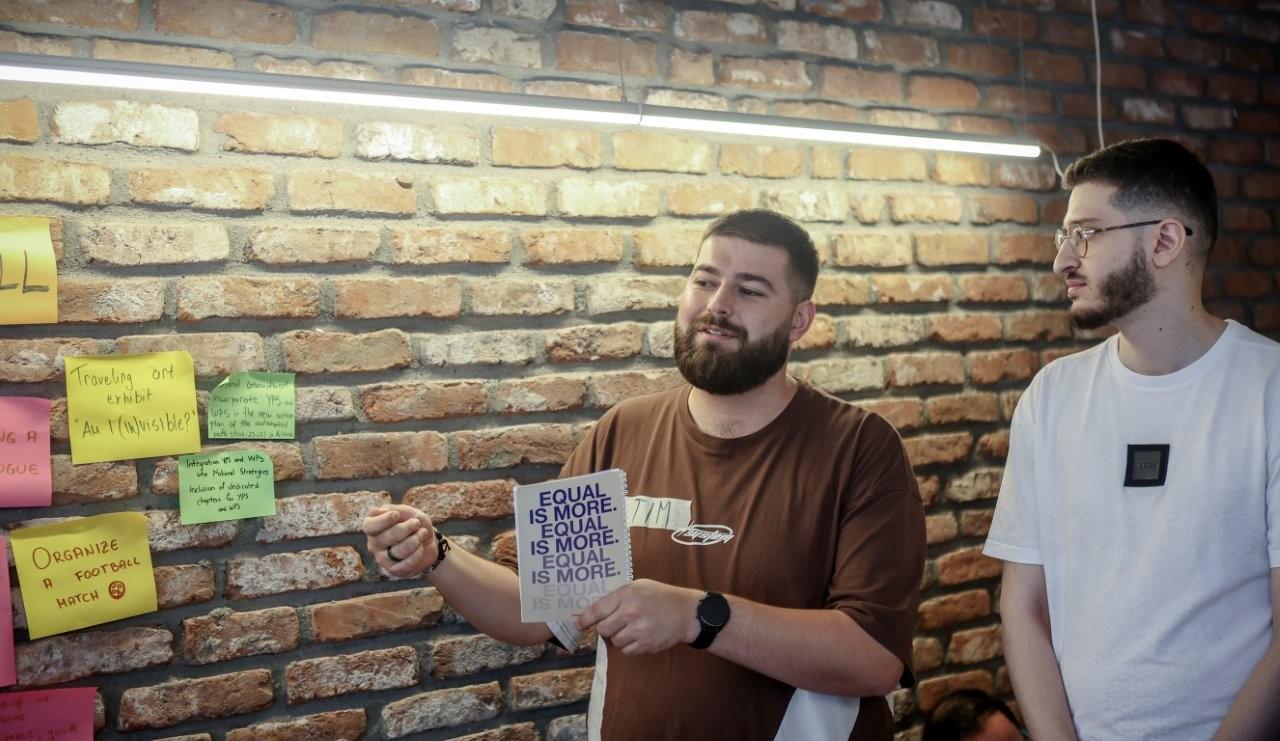 A man with a beard presents while another man listens, surrounded by colorful sticky notes.