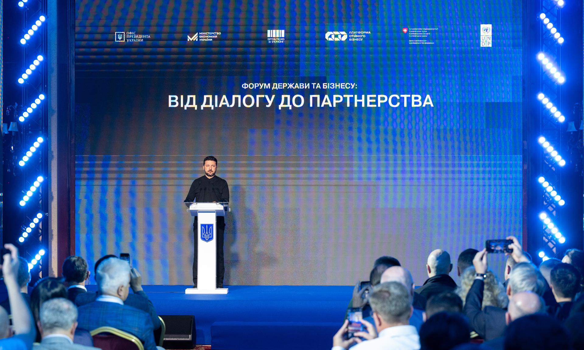 A speaker addresses an audience at a conference, with blue lighting and a backdrop.