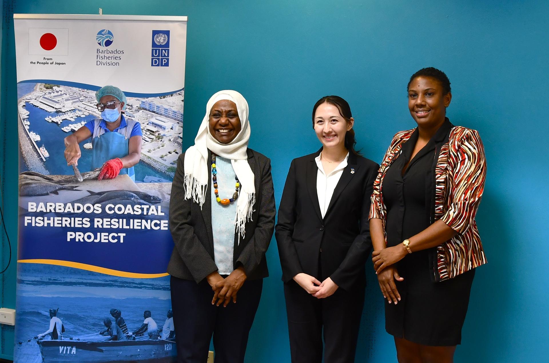 Three women stand together in front of a blue wall, beside a project banner.
