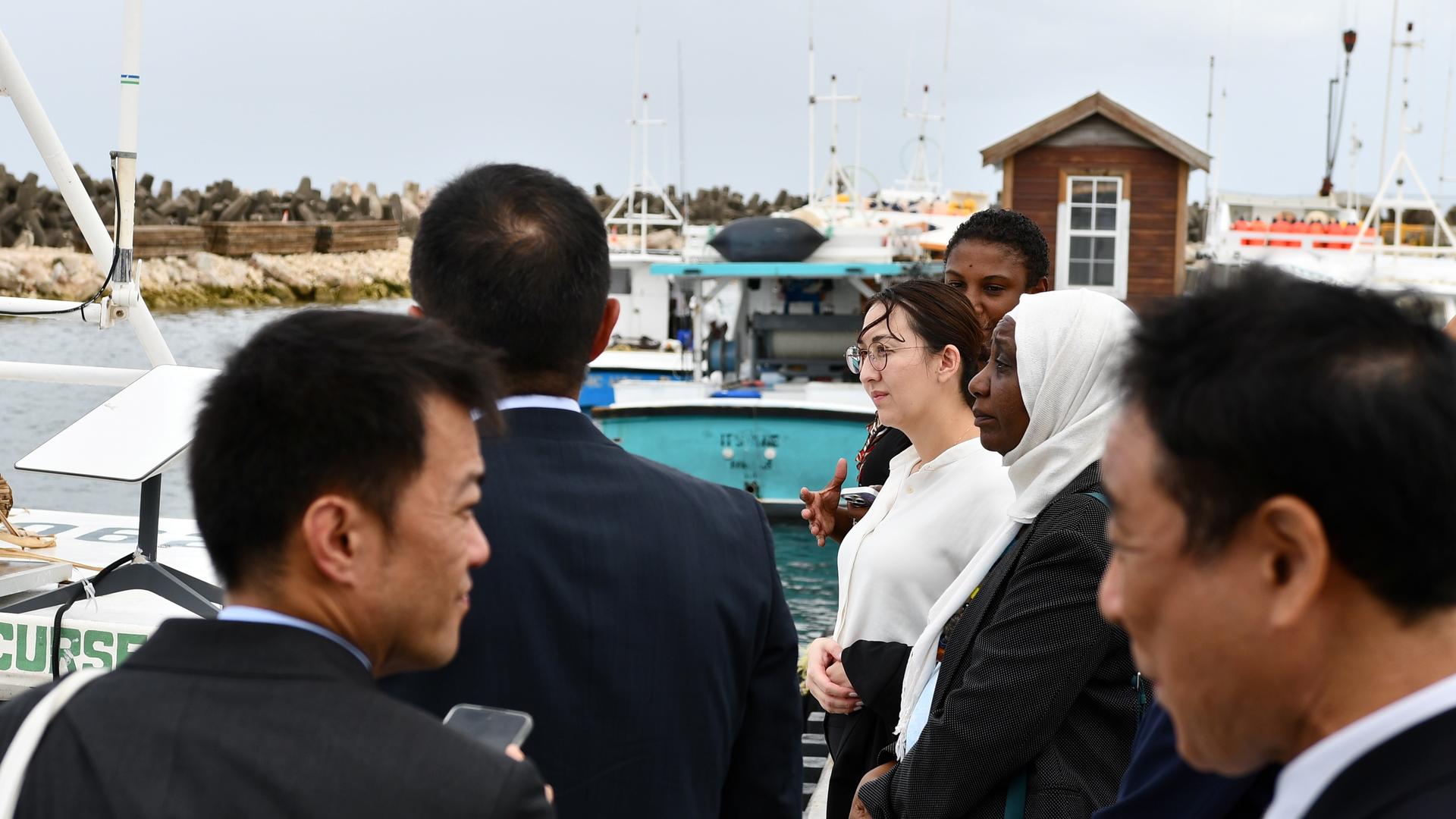 A group of people facing a harbor with boats and a wooden building in the background.