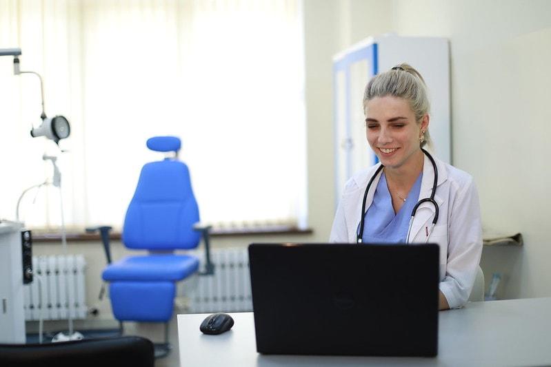 A smiling female doctor works on a laptop in a clinical setting with a blue examination chair.