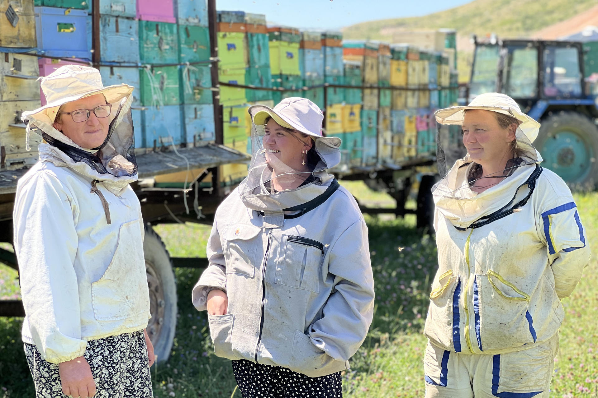 Three beekeepers in protective gear standing in a field, with colorful beehives in the background.