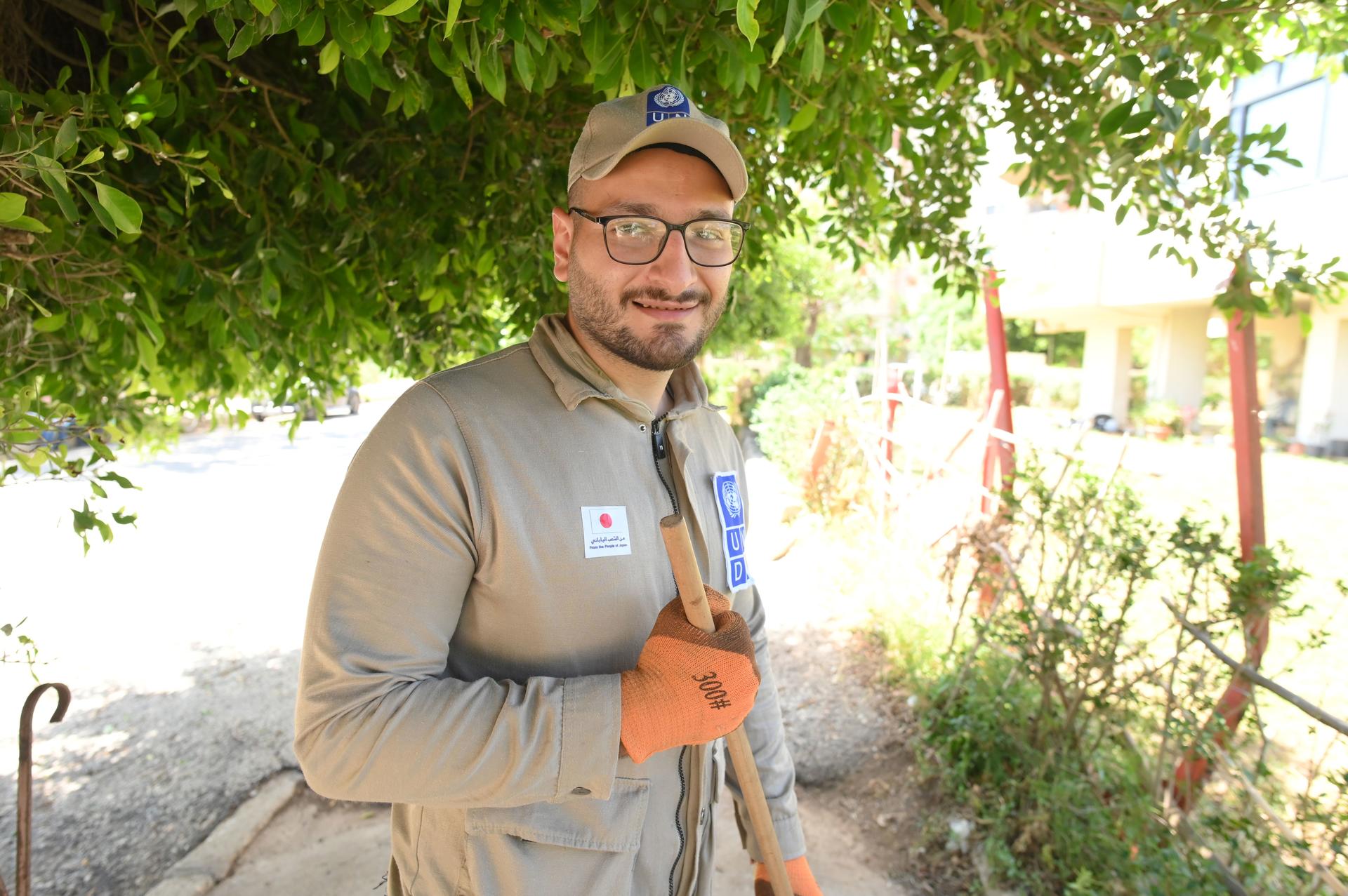 A smiling worker in a beige uniform, holding tools, stands under leafy trees.
