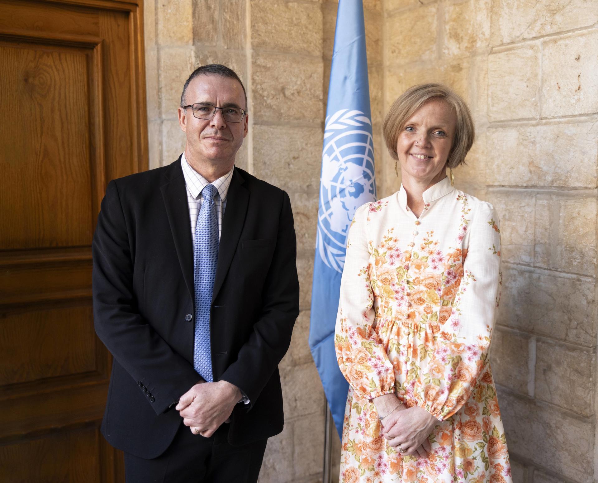 Two individuals stand next to a UN flag in a stone-walled setting, both smiling.