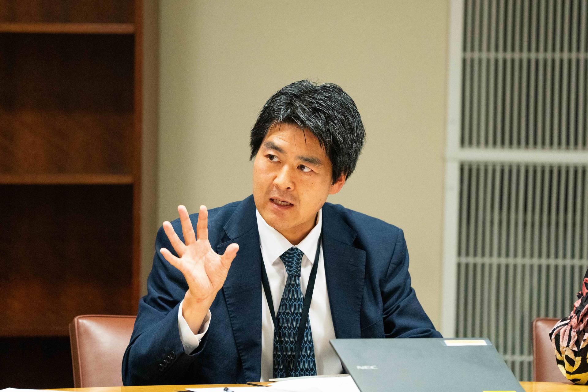 A man in a suit gestures while discussing at a conference table.