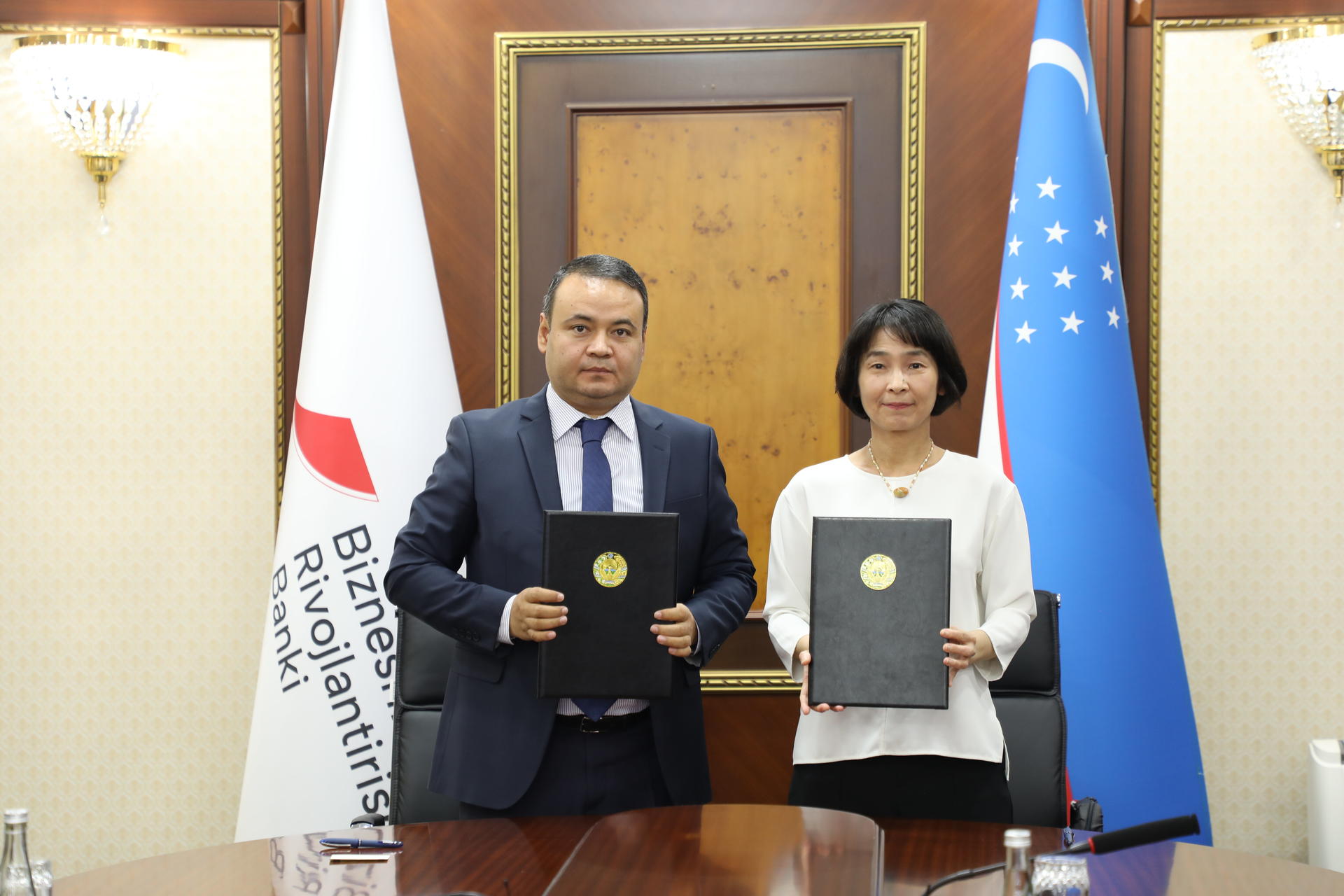 Two individuals holding documents, standing in an office with flags in the background.