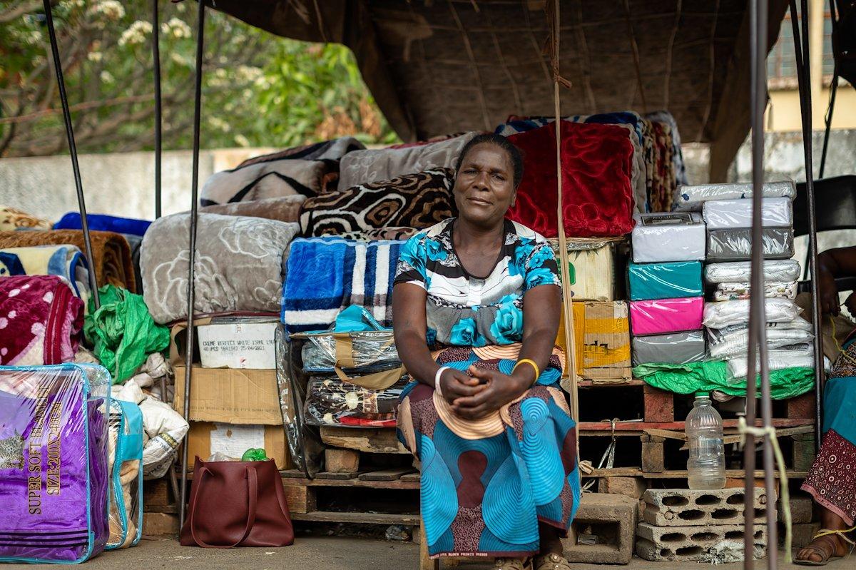 A woman sits in front of colorful blankets and textiles, smiling at her market stall.