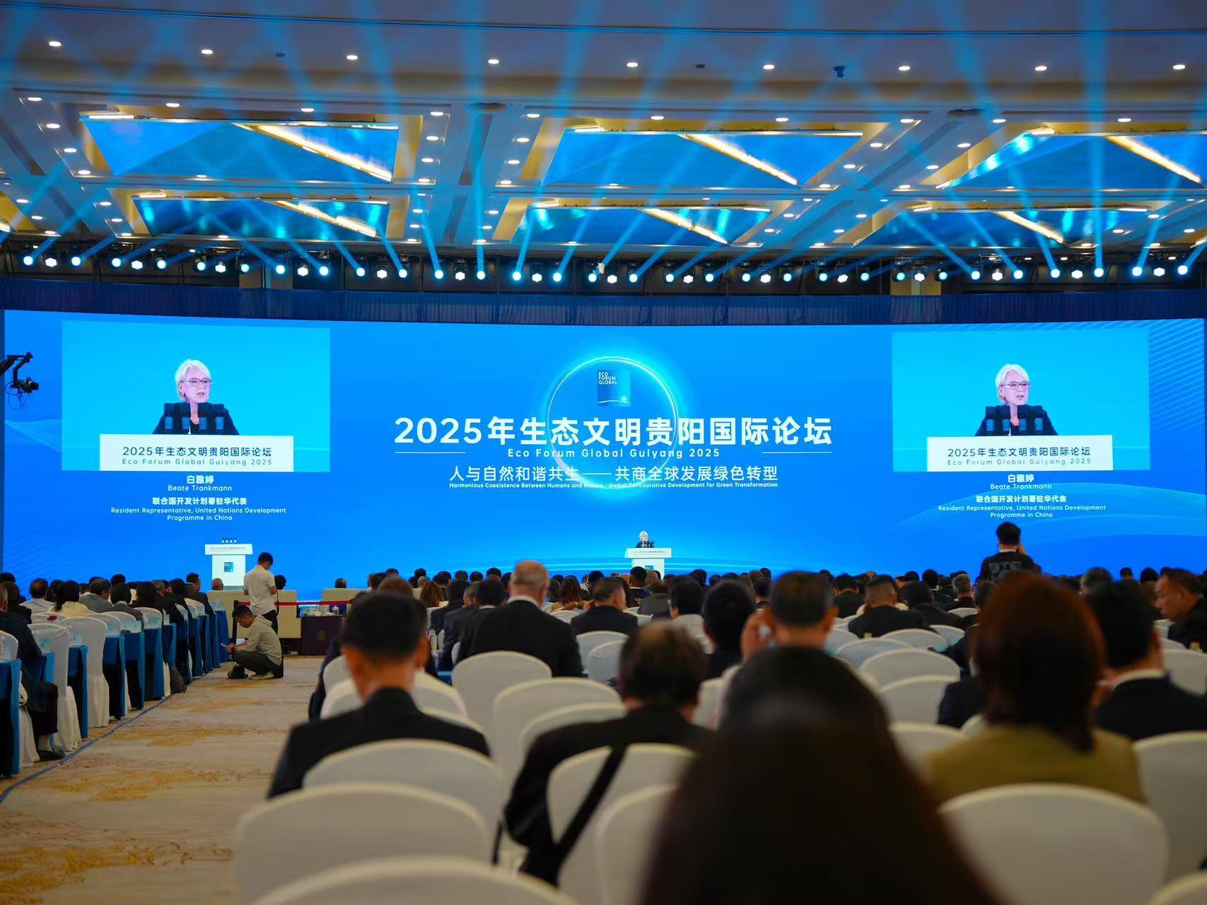 Conference setting with a speaker on stage and an audience in formal attire.