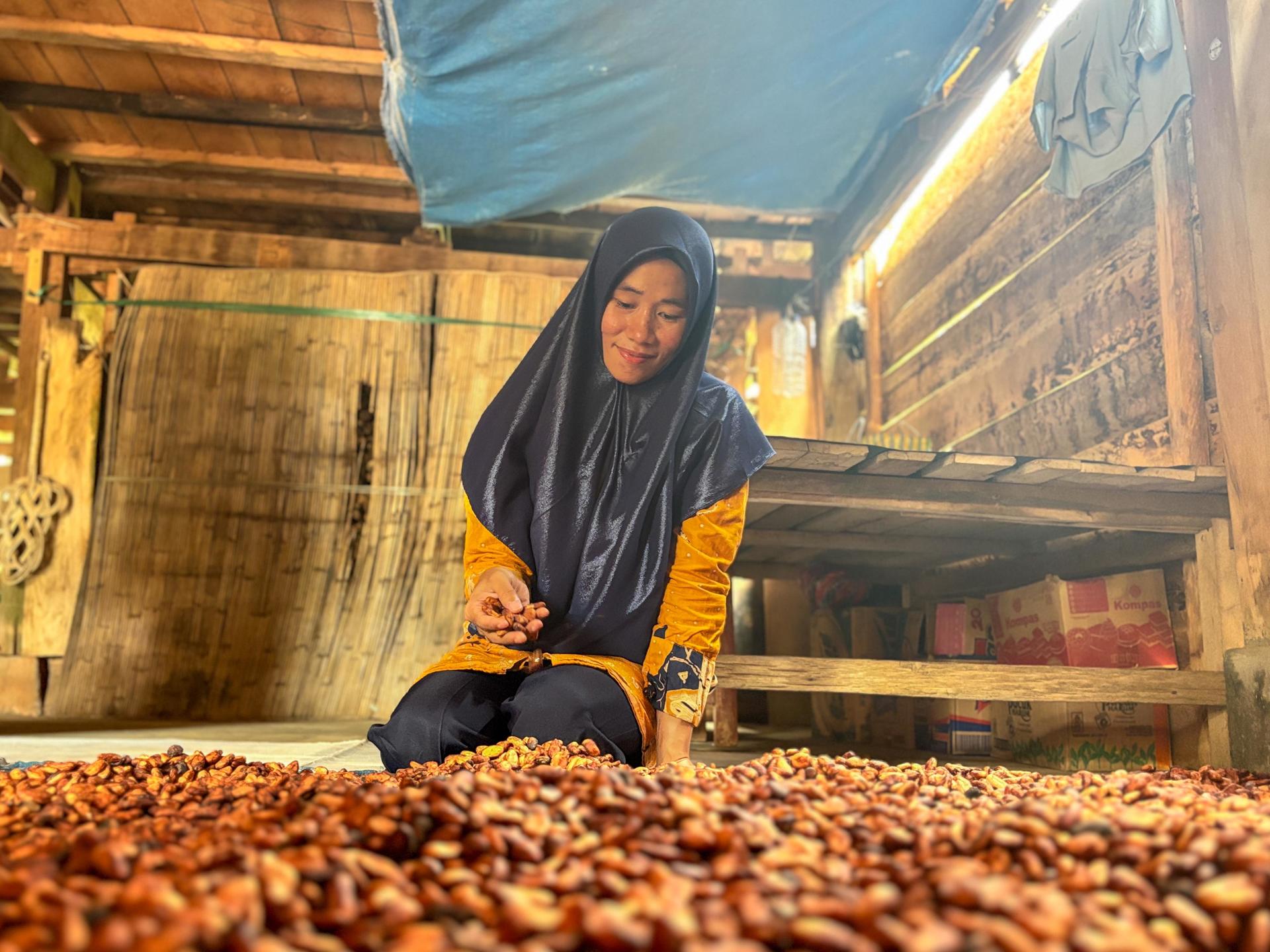 A woman in a dark hijab sorts through brown seeds on the floor of a wooden room.