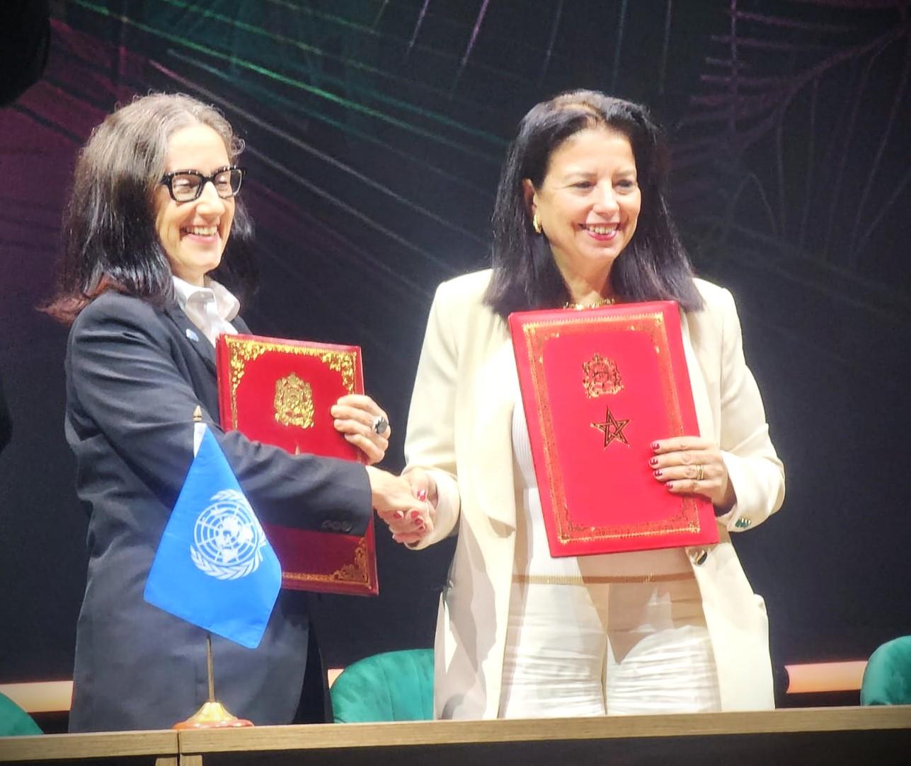 Two women shaking hands while holding red documents, smiling, with a UN flag in the background.