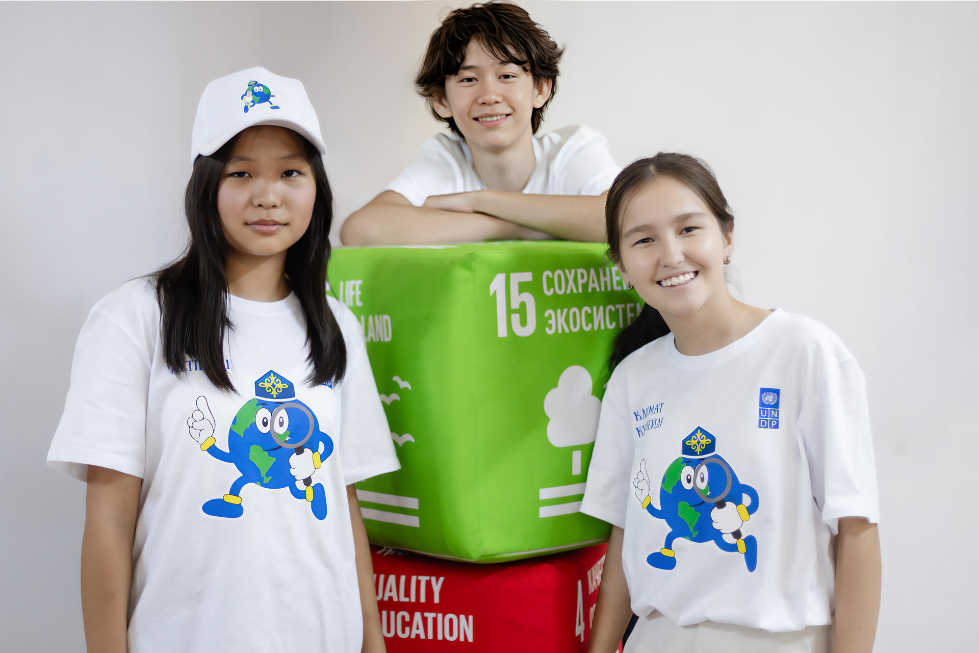 Three young people are smiling, wearing matching shirts and a cap, standing by colorful boxes.