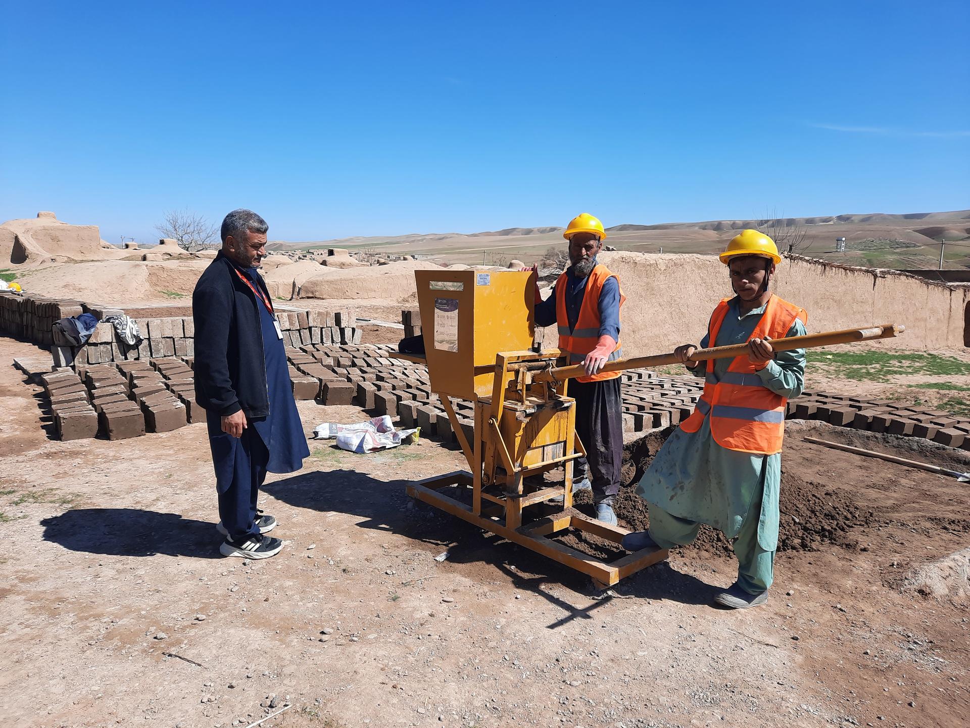 Two workers in hard hats operate machinery on a construction site, with bricks in background.
