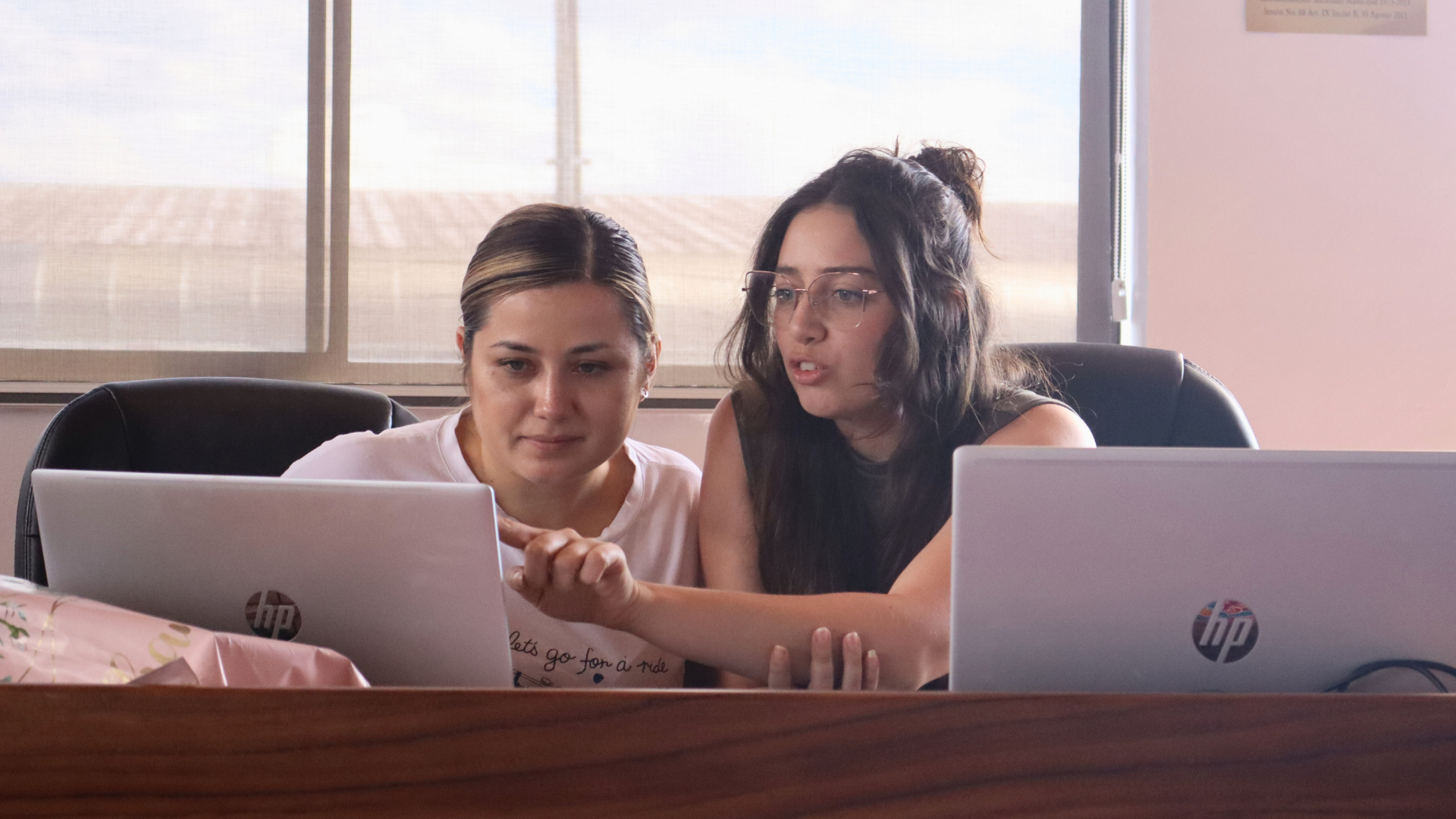 Two women collaborate at a desk with laptops, focused and engaged in conversation.