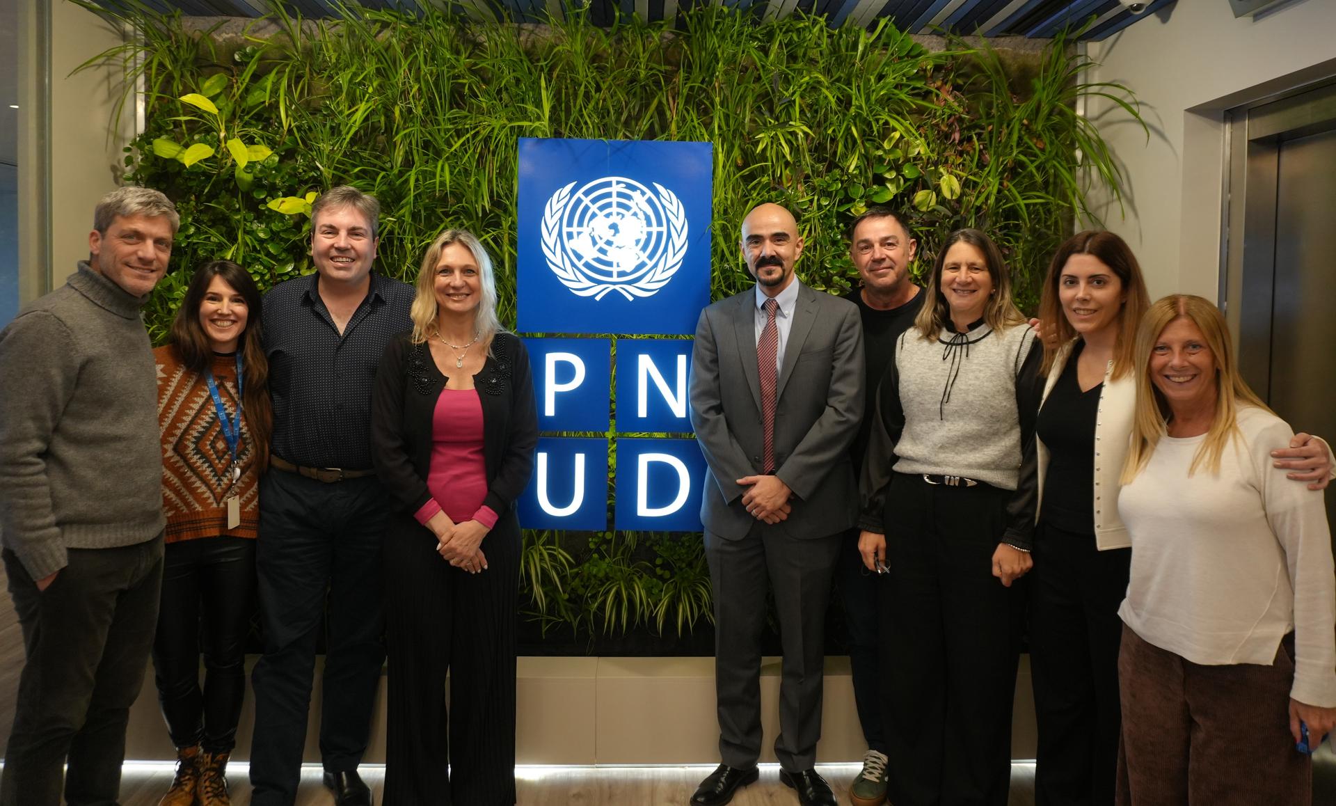 Un grupo de profesionales posando juntos enfrente de una pared con plantas verdes y el logo de PNUD.
