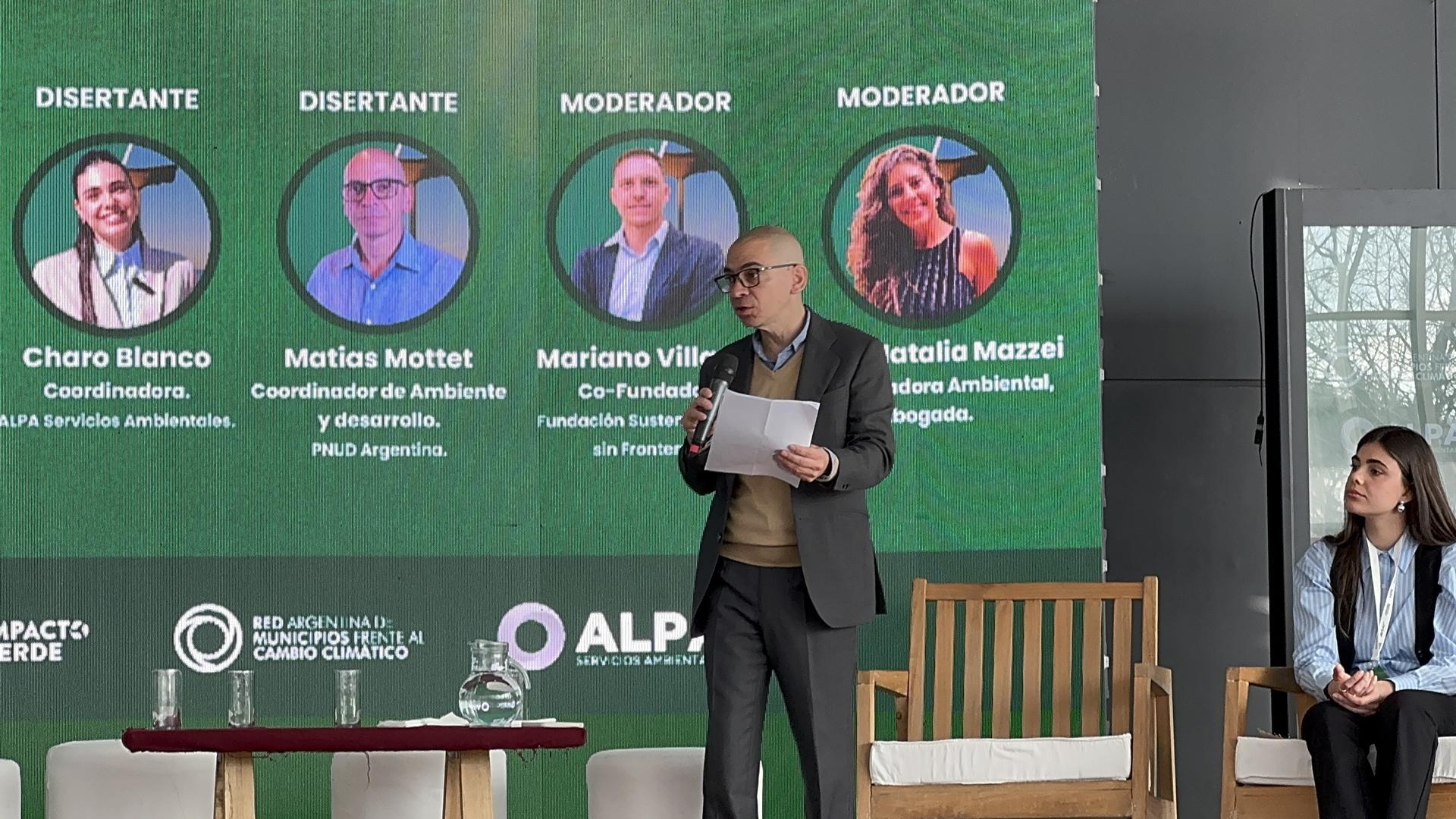 Un orador parado frente a una pantalla con una imagen verde y fotos de panelistas.