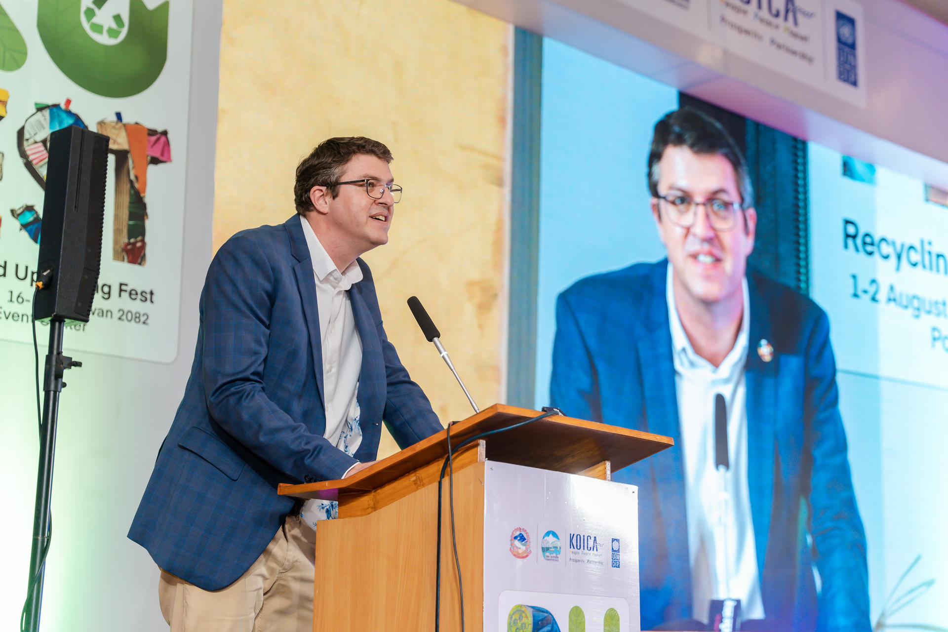 Man in a blue blazer speaks at a podium with a large screen behind displaying his image.