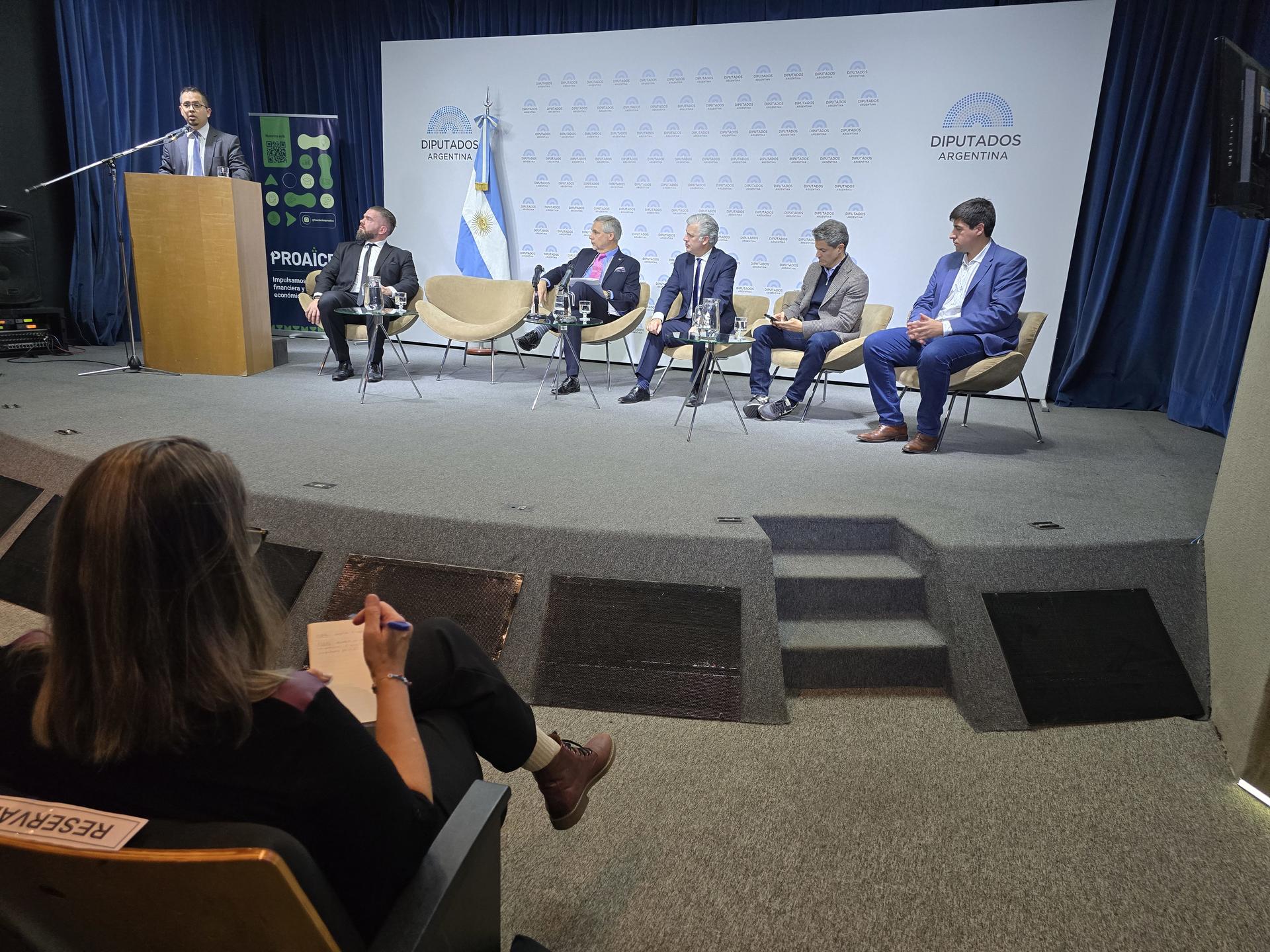 Un panel de discusión con cinco oradores sentados en sillas, con un moderador detrás de un podio.