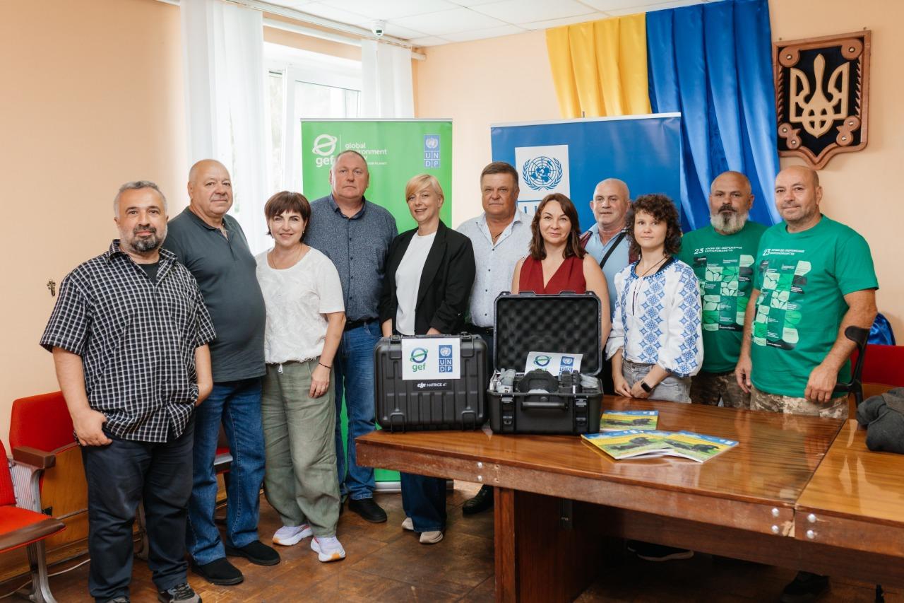 Group of diverse individuals standing together in a room, with equipment on a table and flags in the background.
