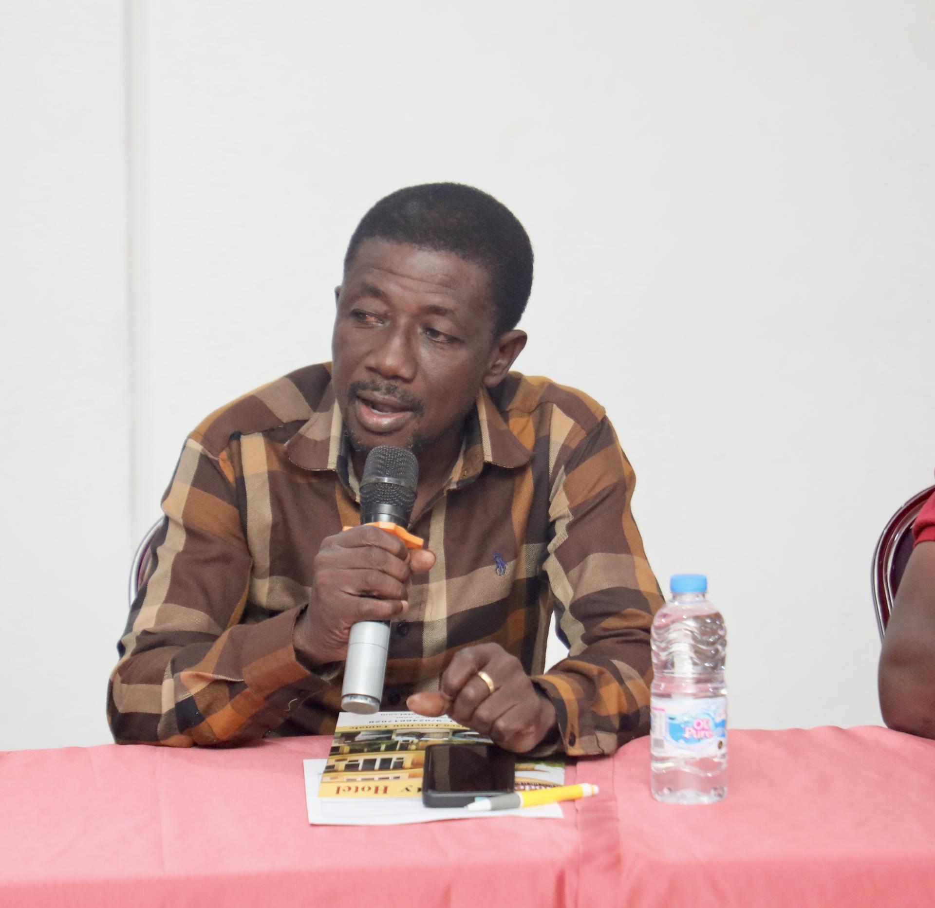 Man speaking into a microphone at a table with a woman and a water bottle.