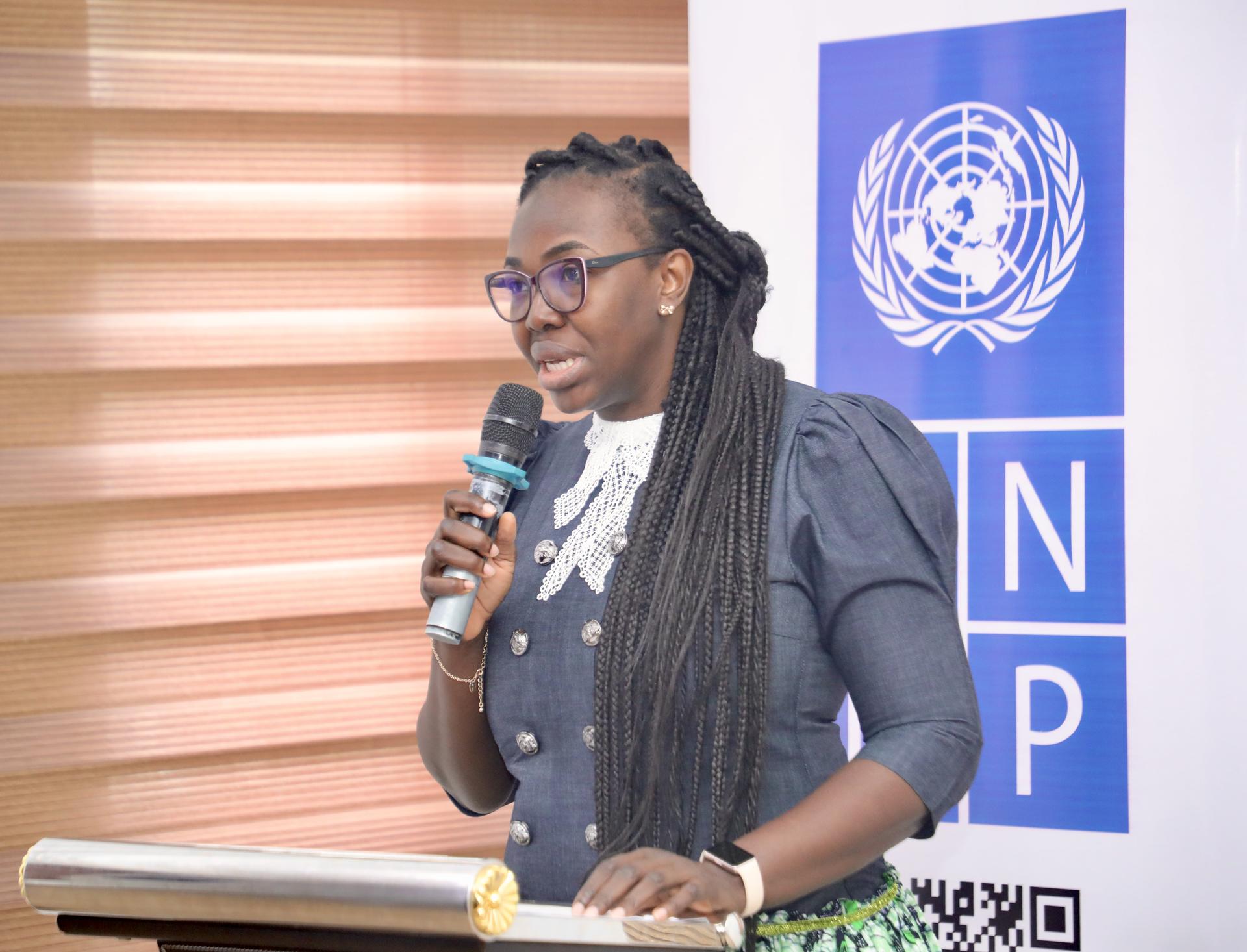 A woman with long braided hair speaks into a microphone at a podium, UN logo in background.