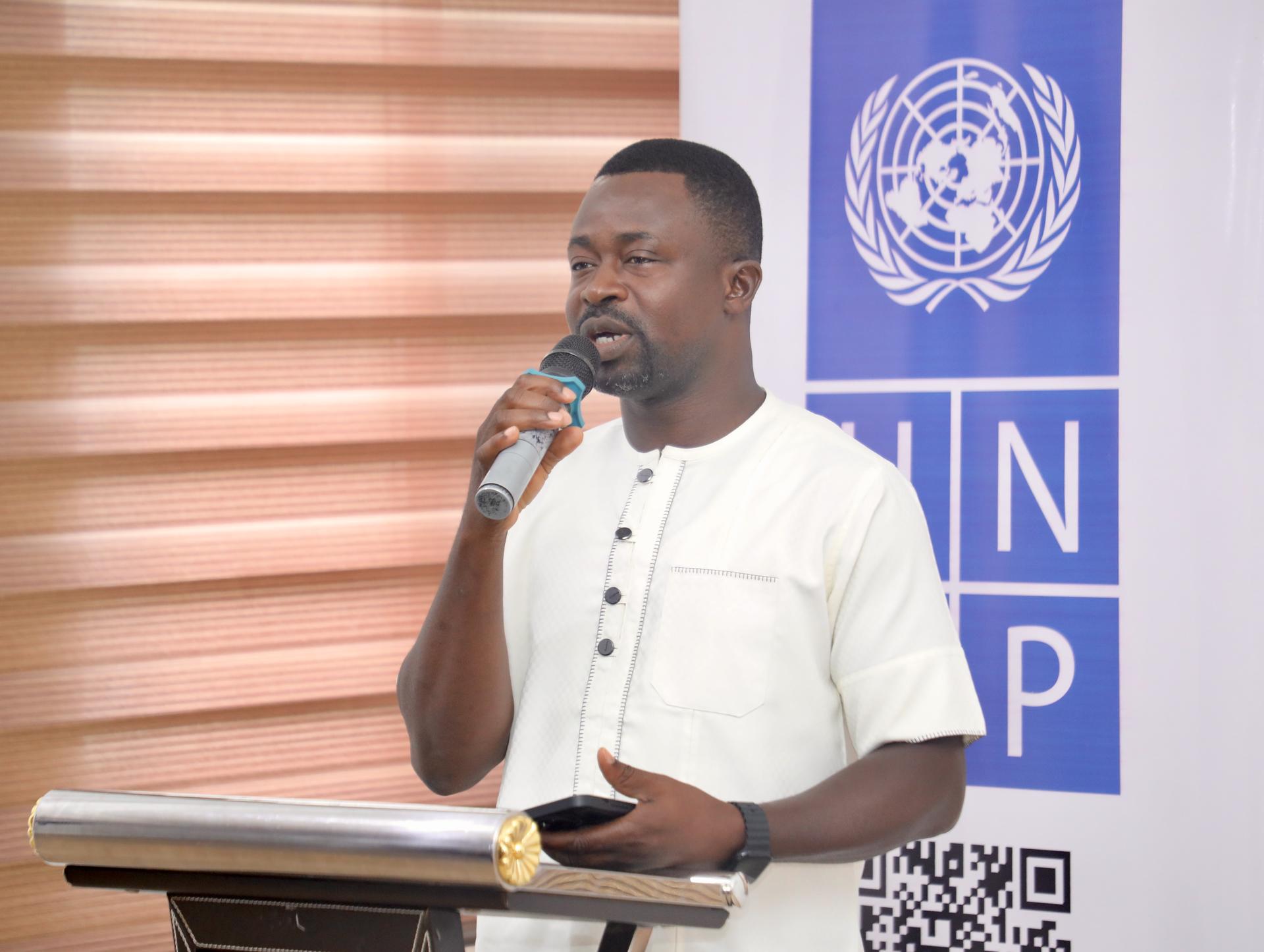 A man in a white shirt speaks into a microphone at a podium beside a UNDP banner.