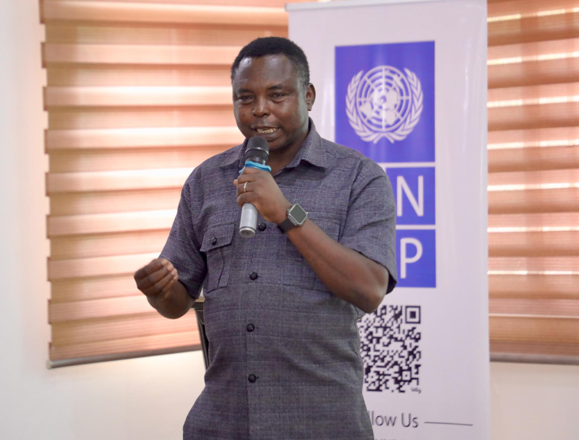 Man speaking into a microphone in a room with blinds, next to a UNDP banner.