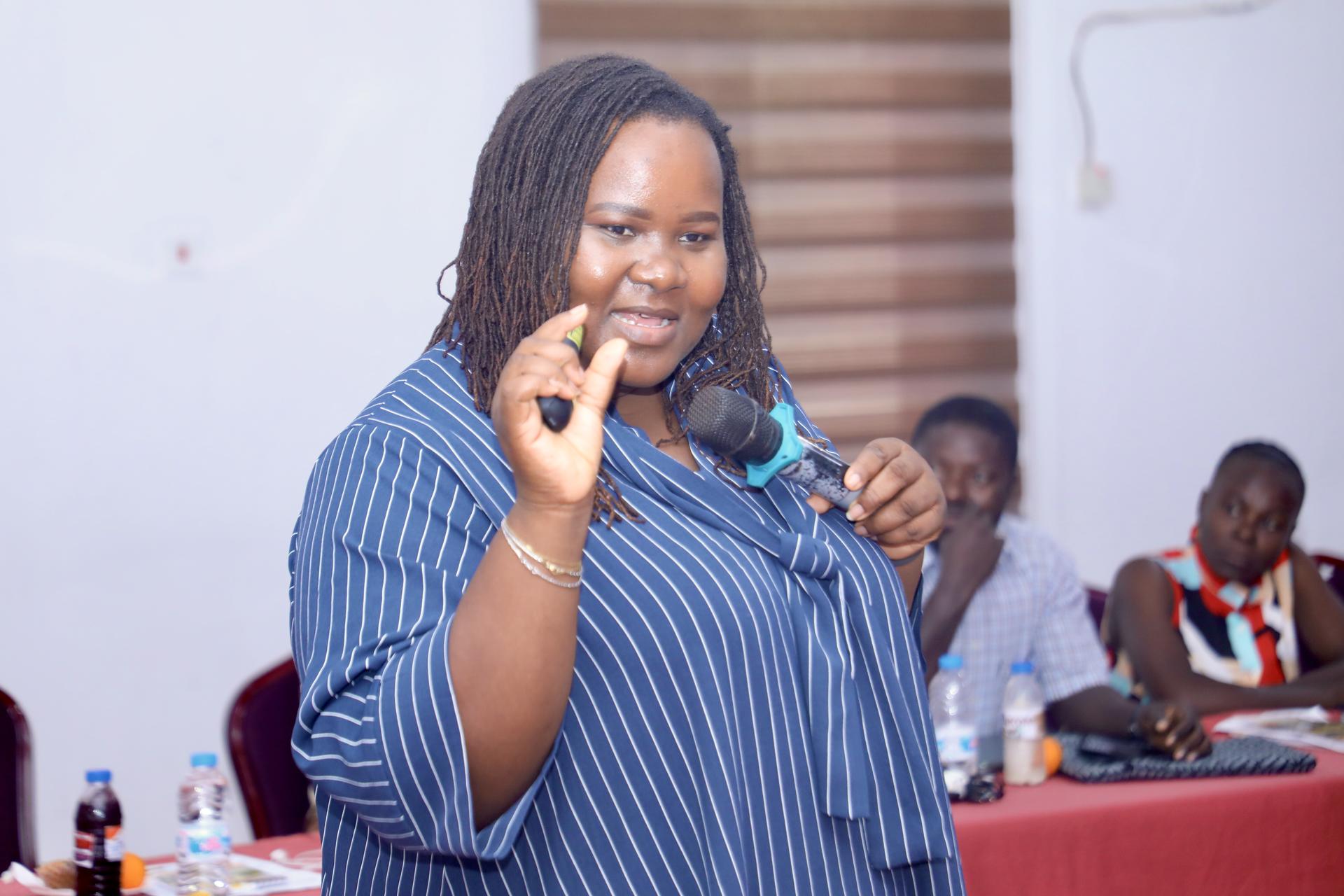 A woman in a striped blue shirt gestures while speaking at a conference, engaged audience in background.