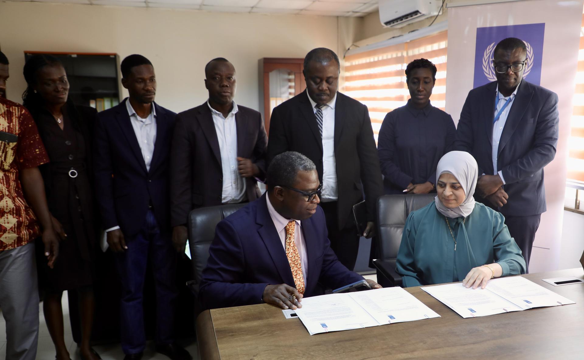 Two individuals seated at a table signing documents, surrounded by a diverse group of colleagues.